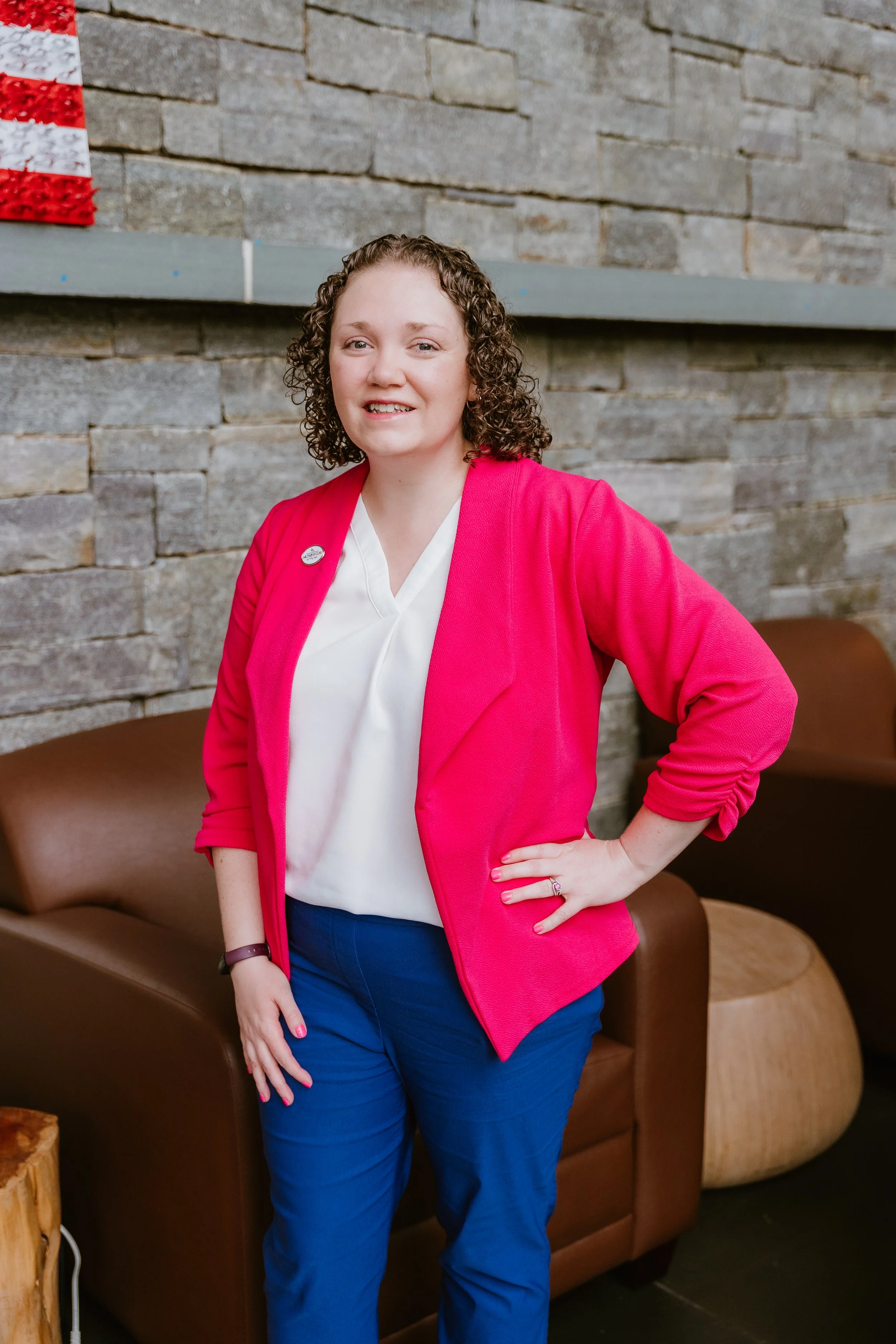 A woman with curly hair wearing a white blouse, bright pink blazer, and blue pants standing indoors against a stone wall and brown leather chairs.
