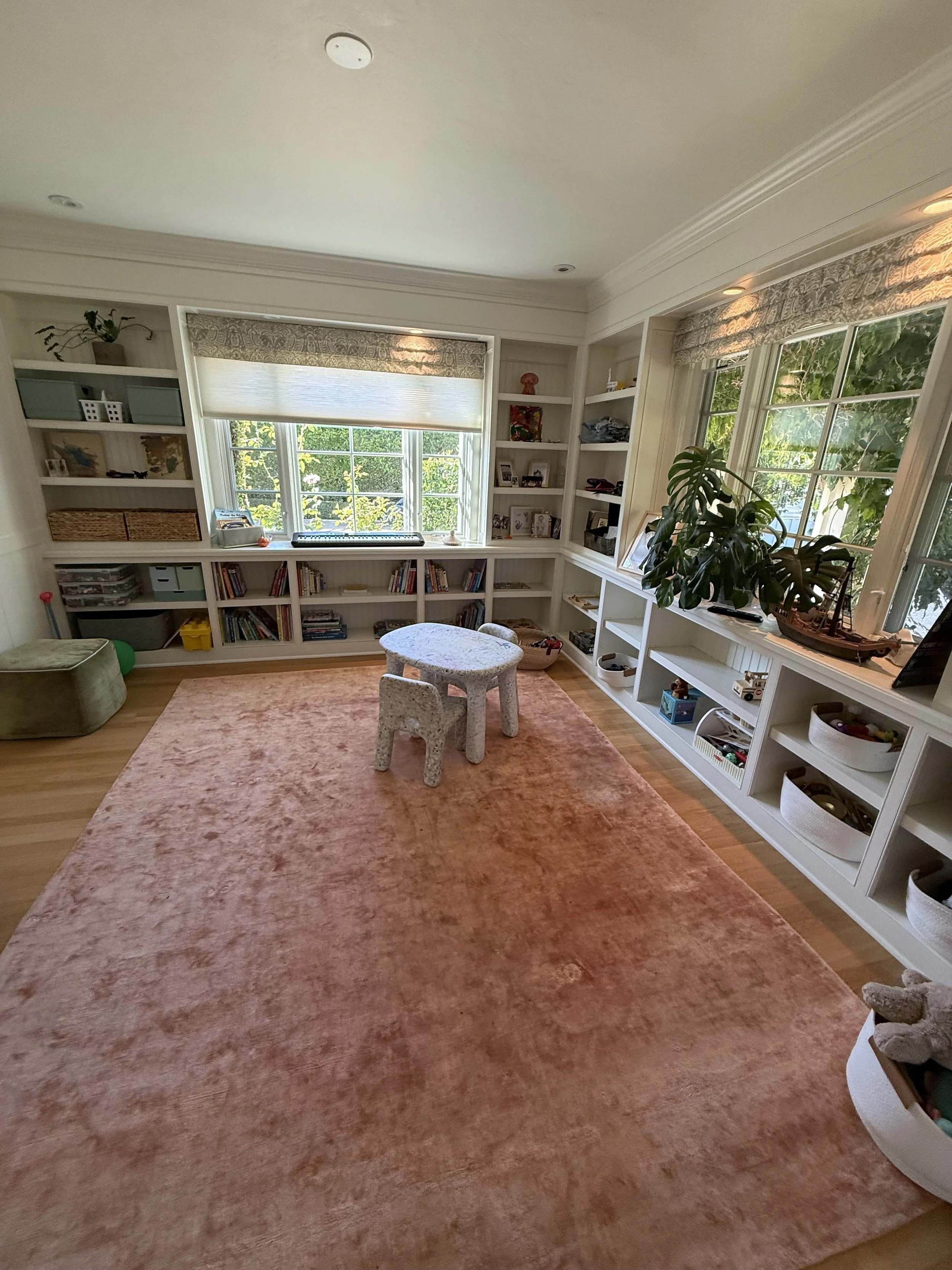 A cozy living room with built-in white bookshelves filled with books, baskets, and decorative items, a large window allowing natural light, a pink rug on wooden flooring, a small table with matching chairs, and houseplants on the window sill and shelves.
