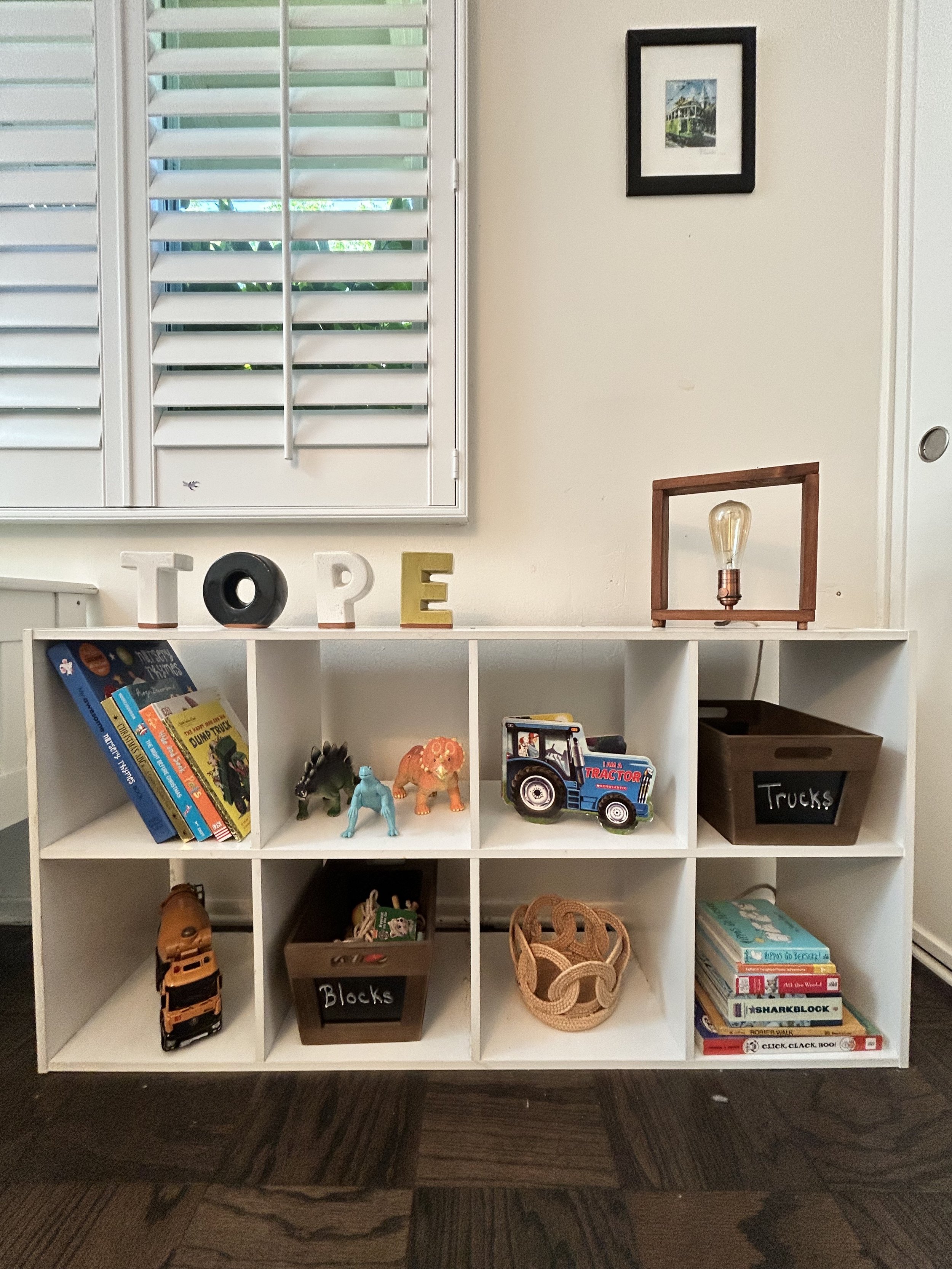 A white cubby shelf with books, toy animals, a toy tractor, and labeled storage bins, with decorative letters spelling 'TOPE' and a framed picture on the wall above.