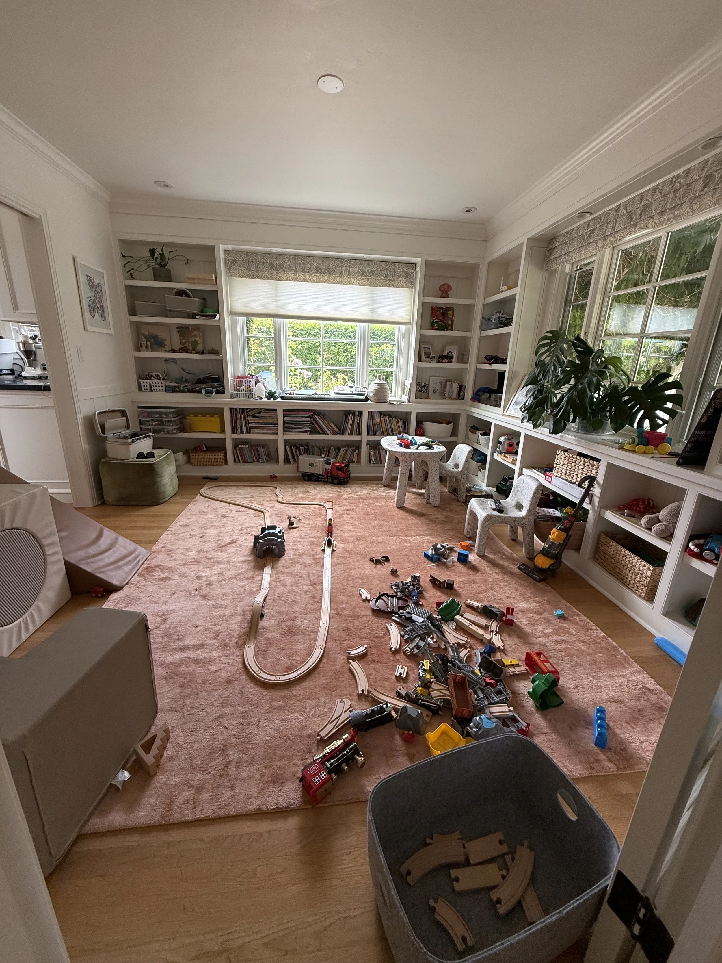 A children's playroom with scattered toy trains, tracks, and small chairs. The room has large windows, built-in shelves filled with books and toys, and a pink area rug over wooden floors.