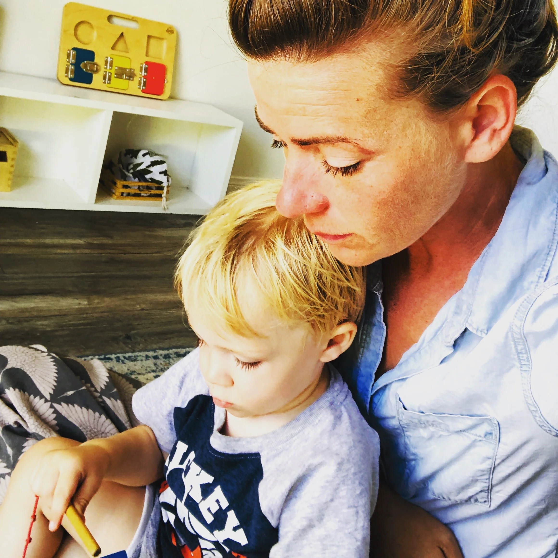 A woman and a young boy sitting closely together, with the woman leaning her head down towards the boy. The boy has blonde hair and is focused on a game or activity, holding a small object in his hand. Behind them is a shelf with toys and educational items.