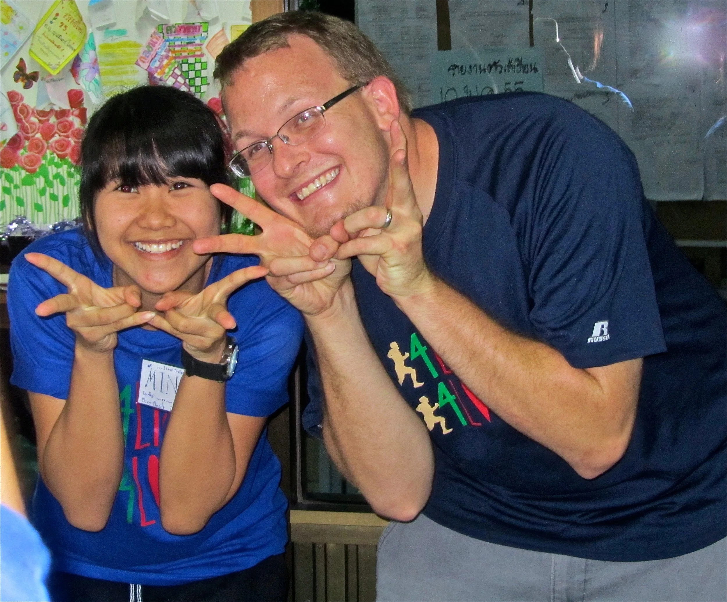 A young woman and a man smiling and making playful hand gestures at the camera, wearing matching blue T-shirts.