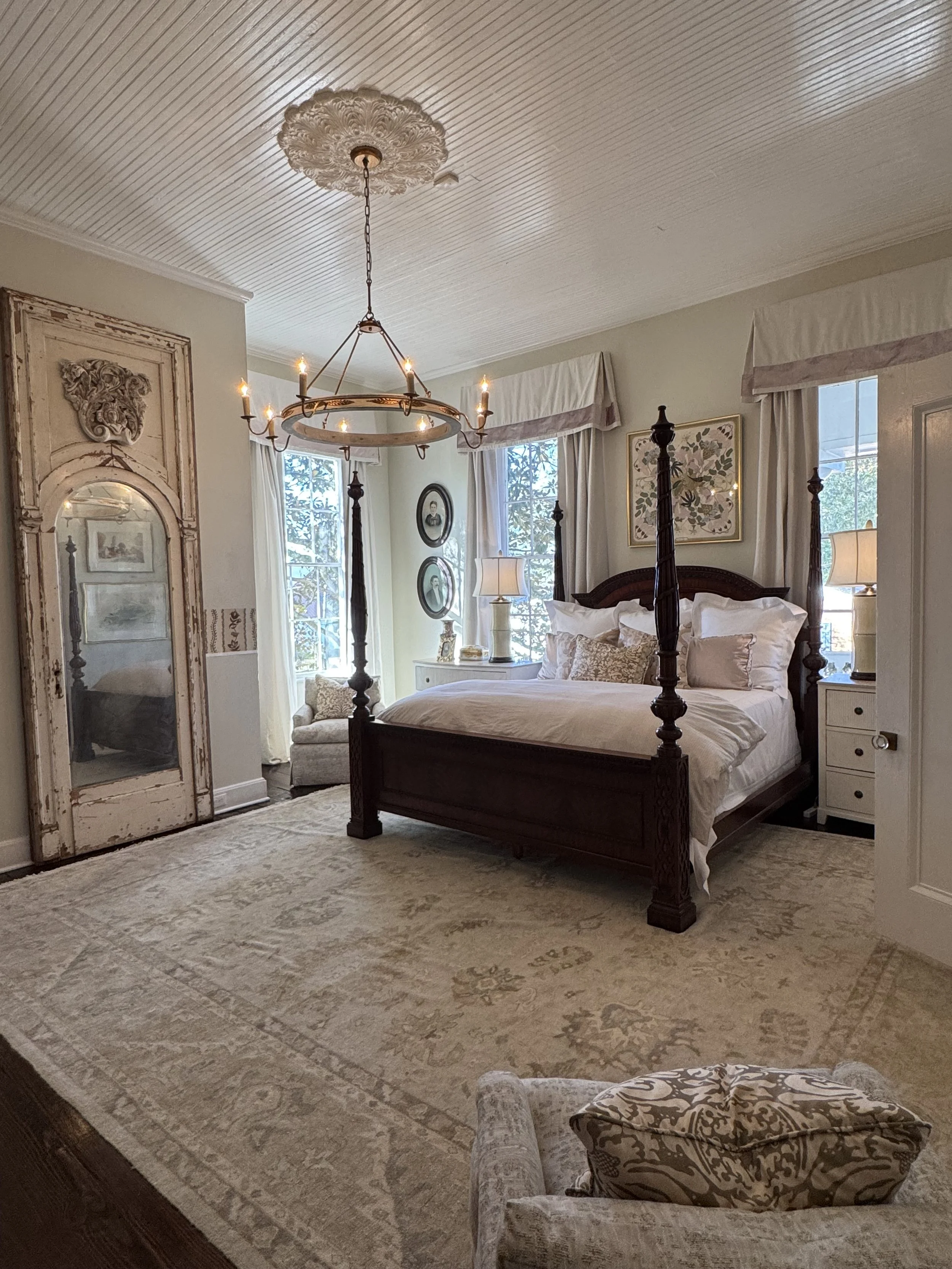 A bedroom with a four-poster bed, a chandelier, two windows with curtains, a vintage mirror, and a chair with a patterned pillow.
