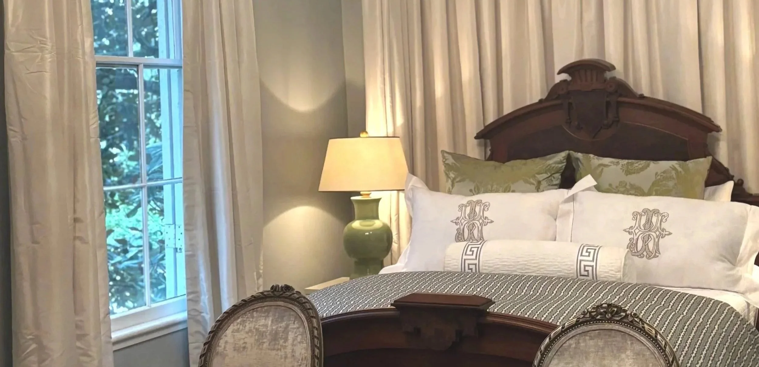 Elegant bedroom with a large wooden headboard, decorative pillows, a bedside lamp, and a window with cream curtains.