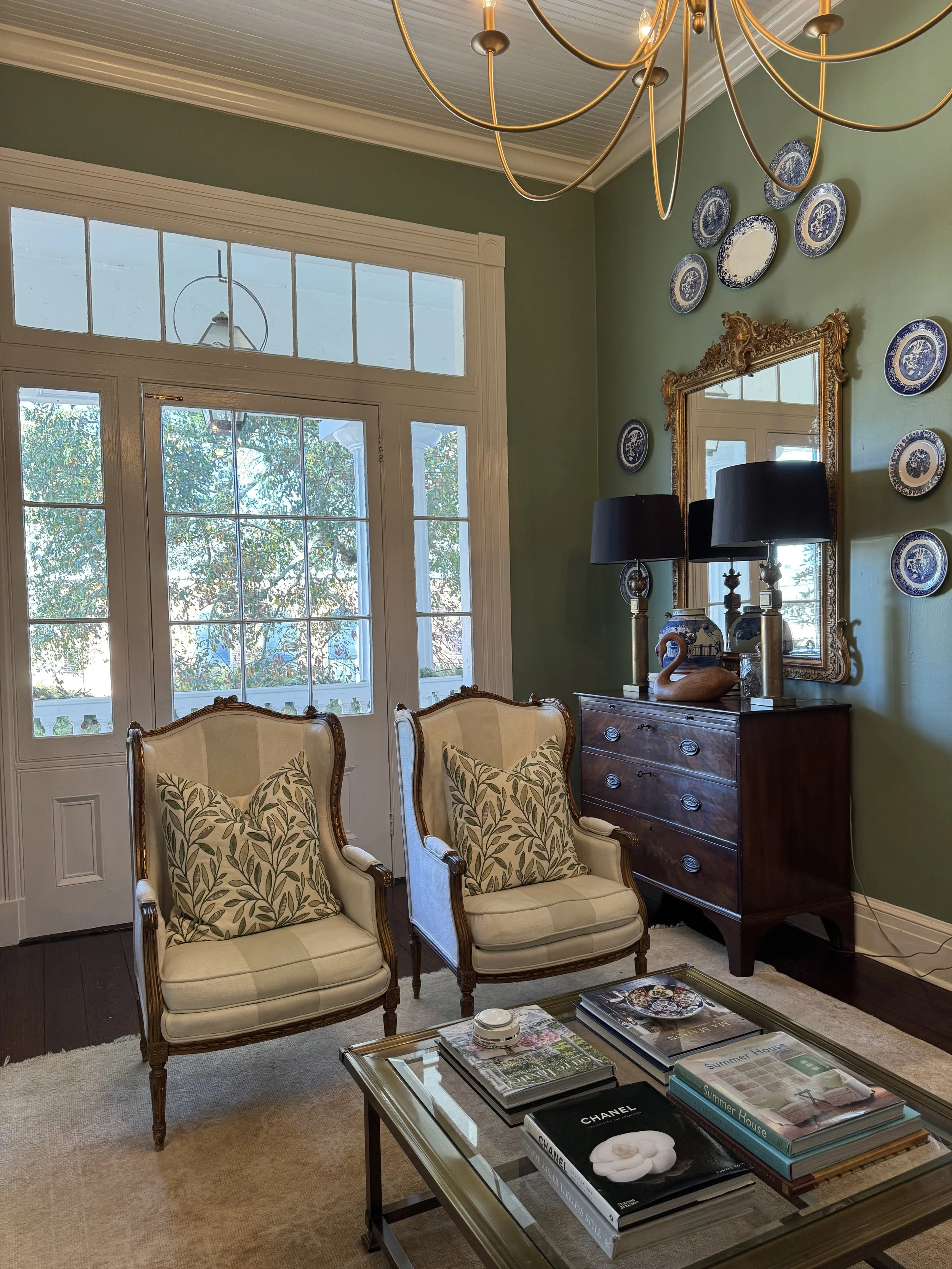 A cozy living room with two vintage armchairs, a glass-top coffee table with books, a dark wood dresser, a large mirror, two black lamps, a gold-framed mirror, and decorative blue and white plates on a green wall.