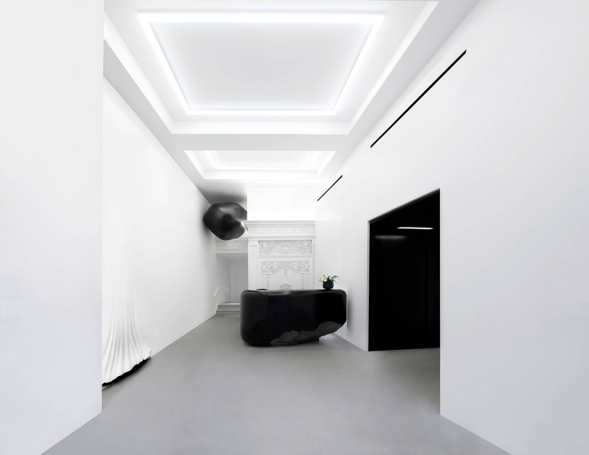 Minimalist white room with a decorative white fireplace, a black abstract sculpture on top of a black piece of furniture, and a black wall niche. The ceiling has recessed lighting panels, and the floor is covered with light gray carpet.
