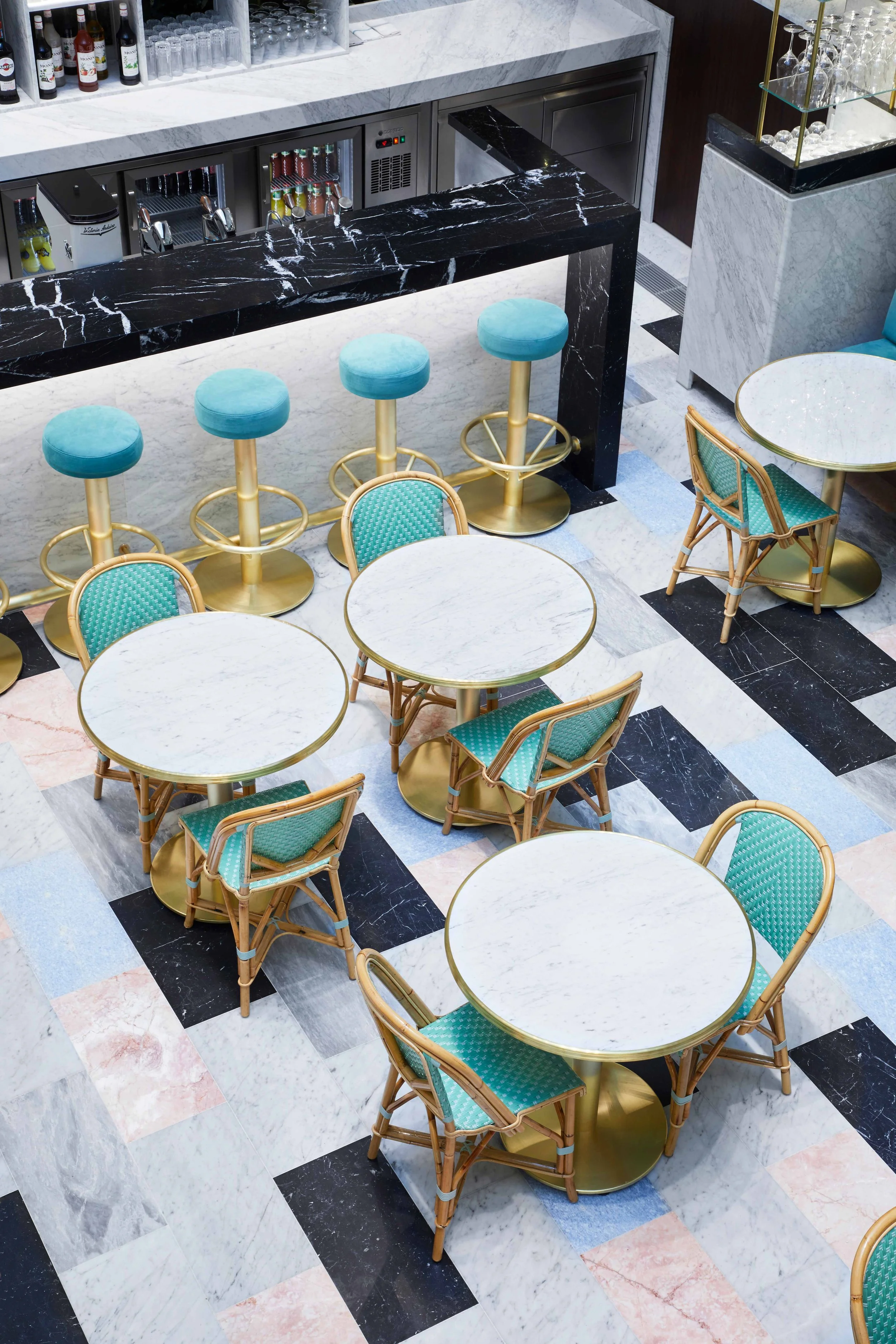 Elegant bar and seating area with white marble tables, turquoise upholstered chairs with rattan frames, and a black marble bar counter with blue barstools featuring gold bases in a stylish setting.
