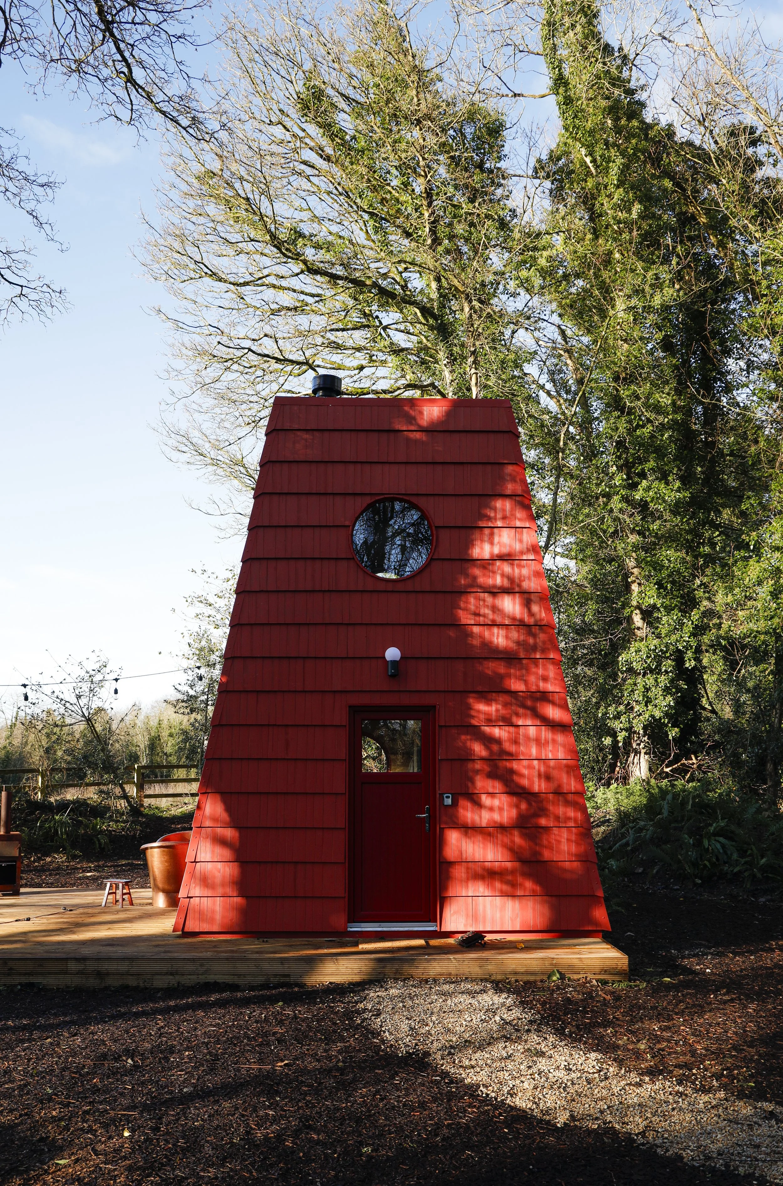 A small red house with a slanted, shingled roof, a circular window near the top, a door with a window, and an outdoor lighting fixture, surrounded by trees.