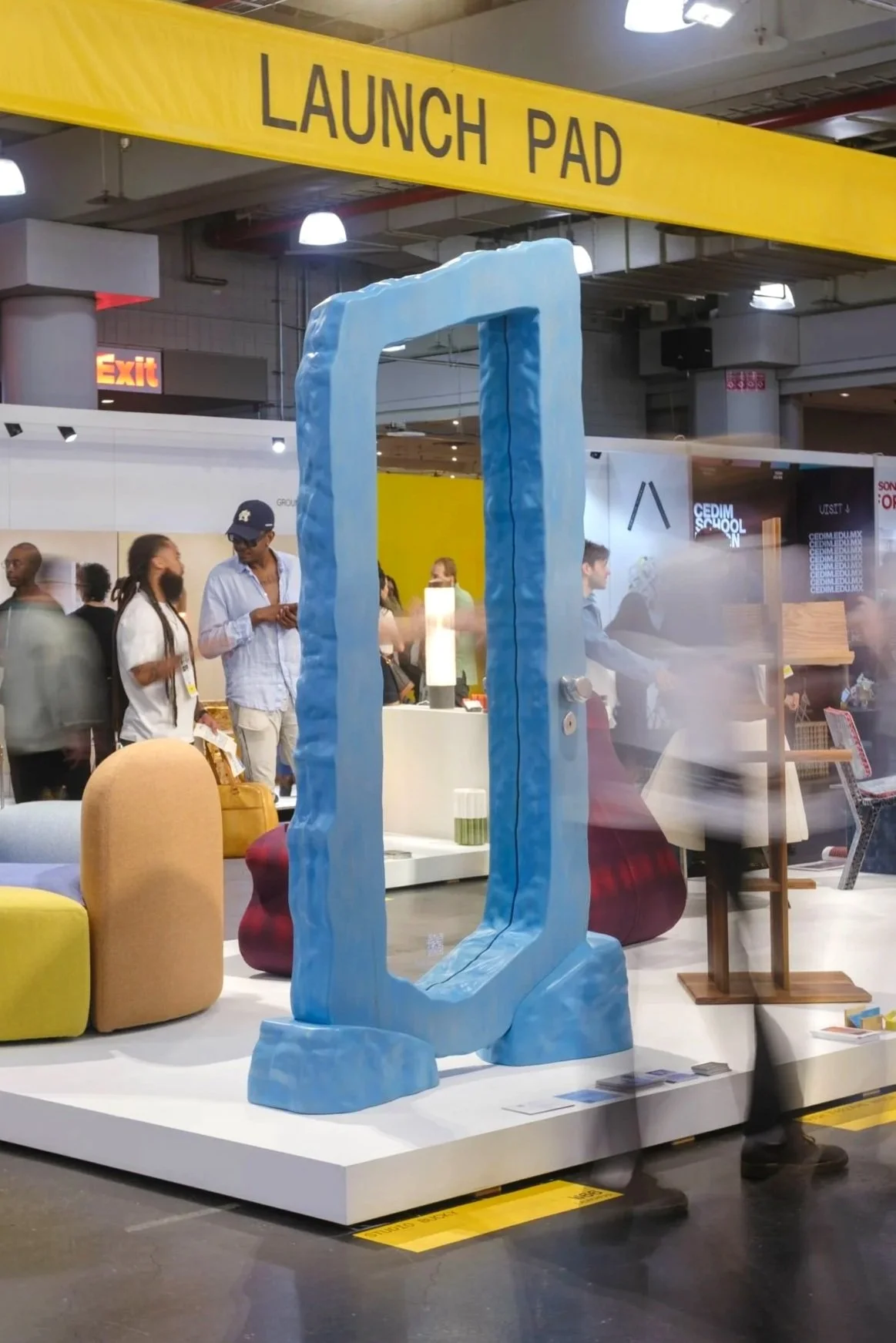 A large blue mirror with irregular edges displayed in an indoor exhibition or store, with people walking and browsing in the background.