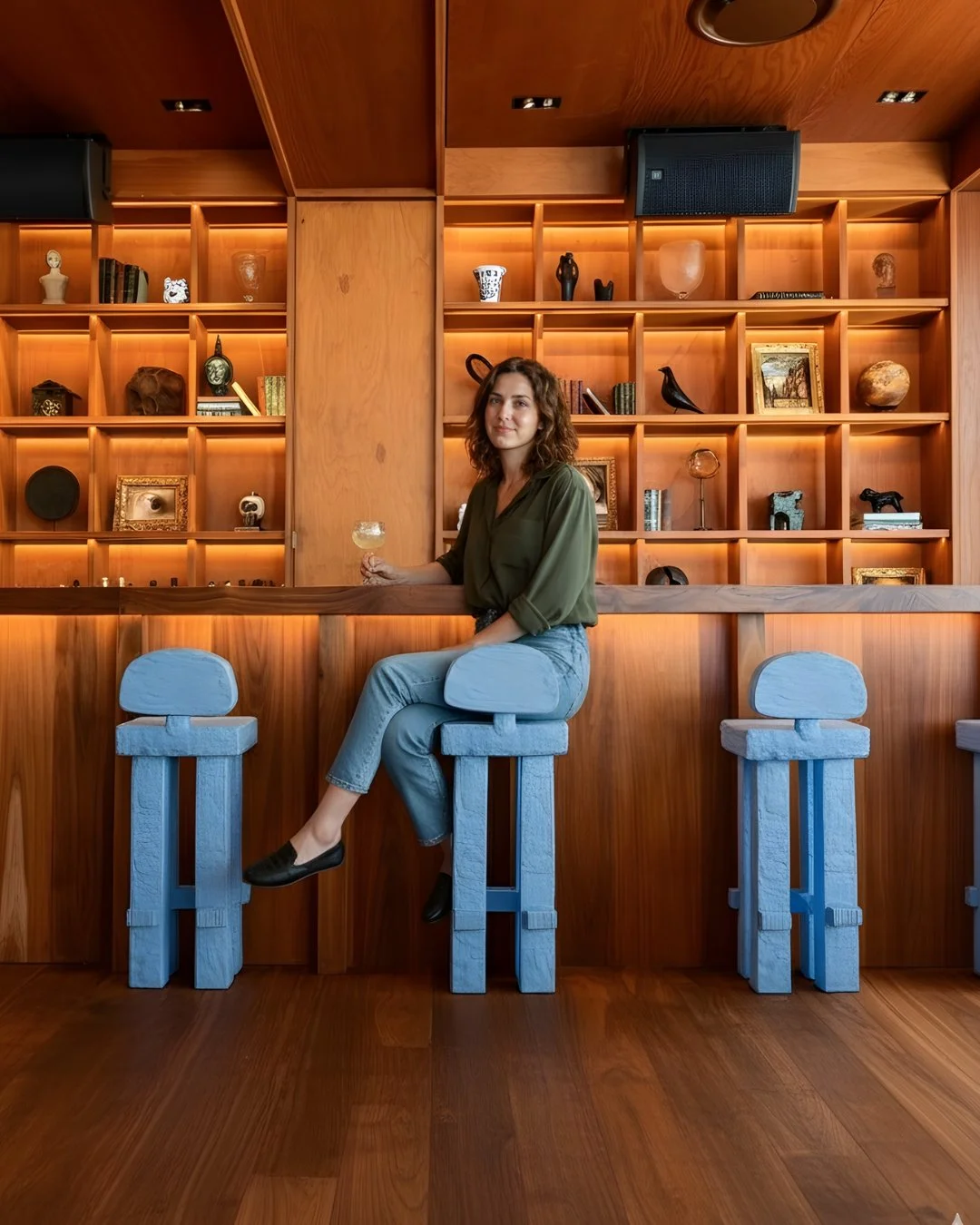 A woman with curly hair sitting on a blue barstool at a wooden bar, holding a drink, with a wooden bookshelf filled with decorative items and art behind her.