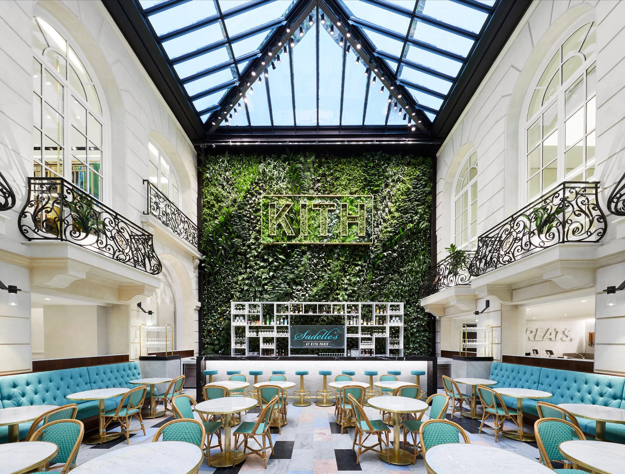 Interior of a stylish cafe or restaurant with a glass ceiling, a lush green wall with a neon sign saying 'KITH', and seating with blue and green upholstery and marble tables.
