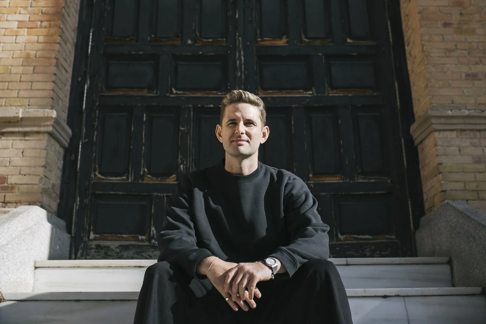 A young man wearing a black sweatshirt and black pants sitting on steps in front of a large black door with brick walls on either side.