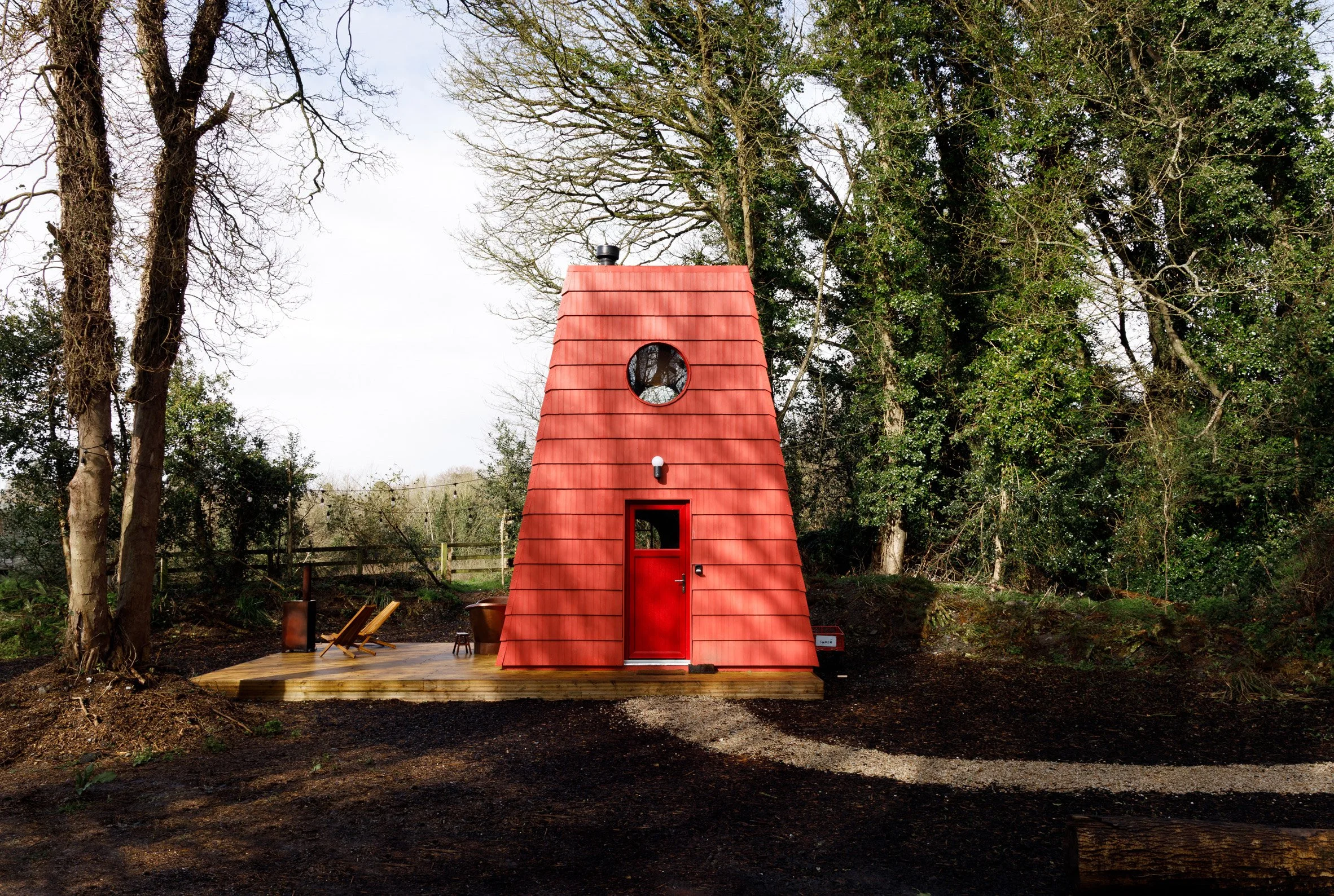 A small red house with a slanted, shingled roof, a circular window near the top, a door with a window, and an outdoor lighting fixture, surrounded by trees.