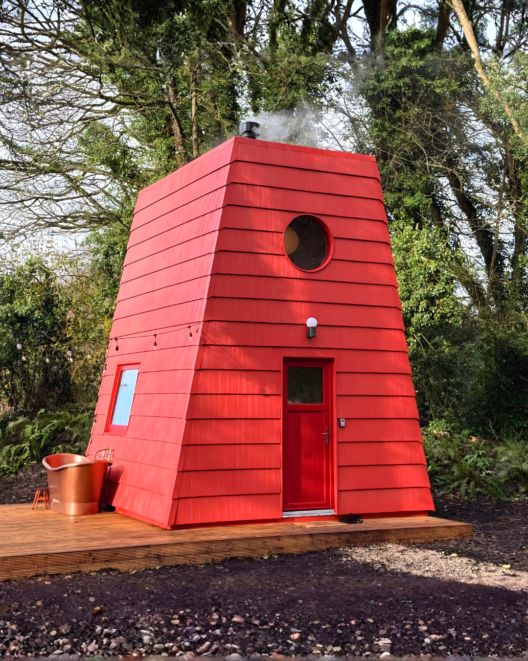 A small red house with a slanted, shingled roof, a circular window near the top, a door with a window, and an outdoor lighting fixture, surrounded by trees.