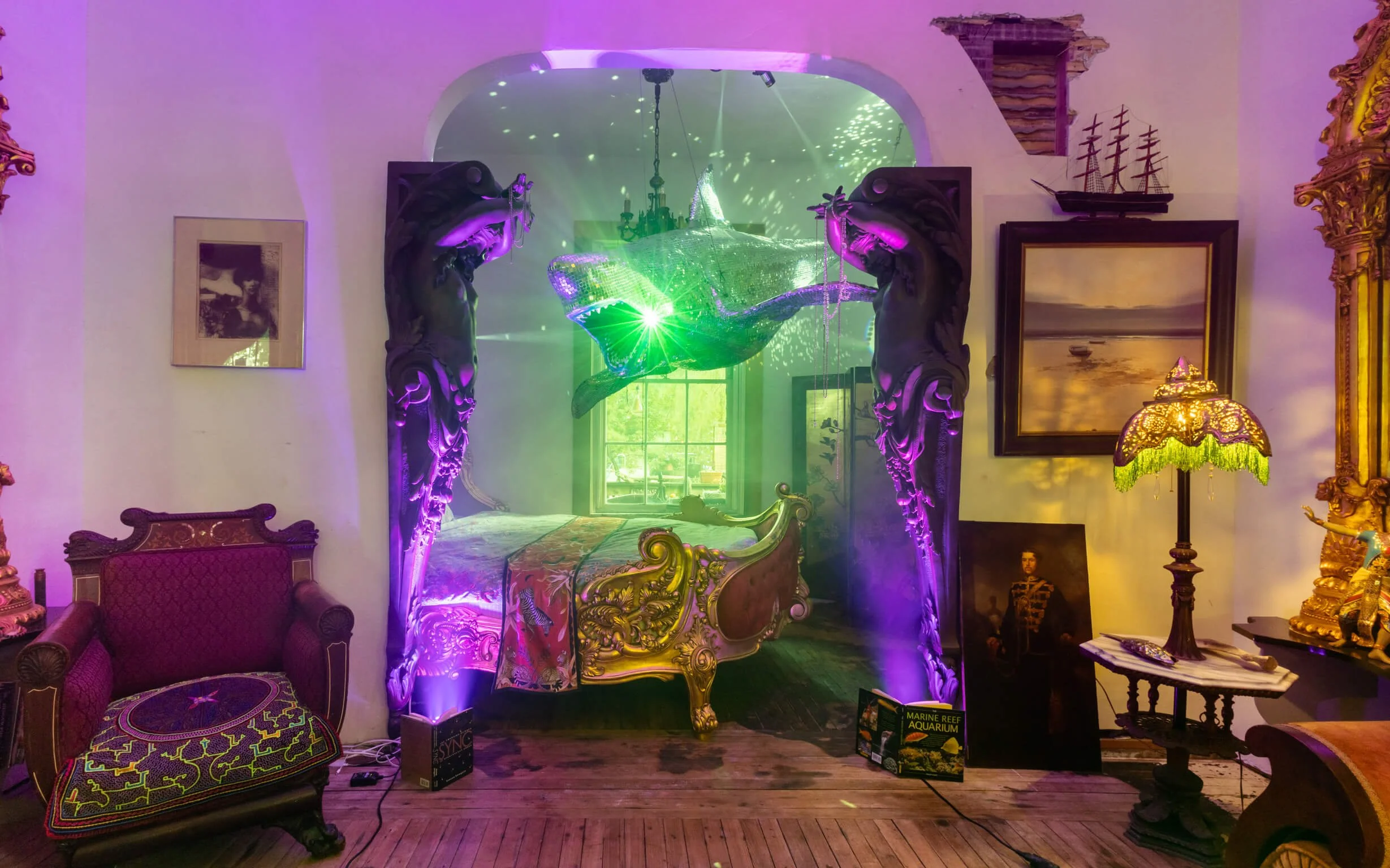 Decorative bedroom with ornate bed, illuminated by purple and green lights, featuring a large fish lantern suspended above, and surrounded by vintage furniture, art, and a colorful lamp.