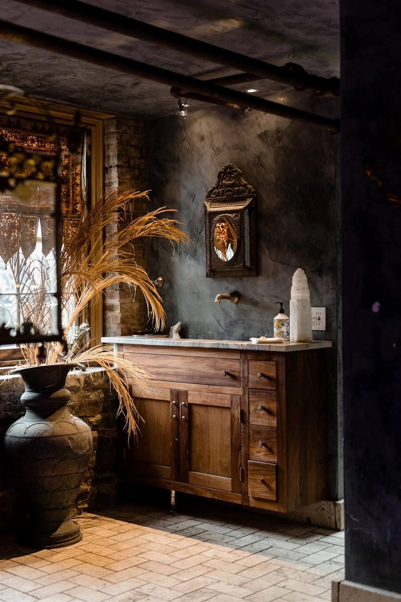 Rustic bathroom corner with wooden vanity, marble countertop, ornate mirror, decorative items, and dried plants near a window with lace curtains.