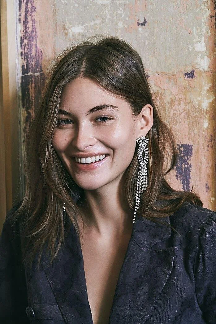A woman with shoulder-length brown hair wearing large sparkly earrings and a dark textured jacket, smiling at the camera against a rustic background.