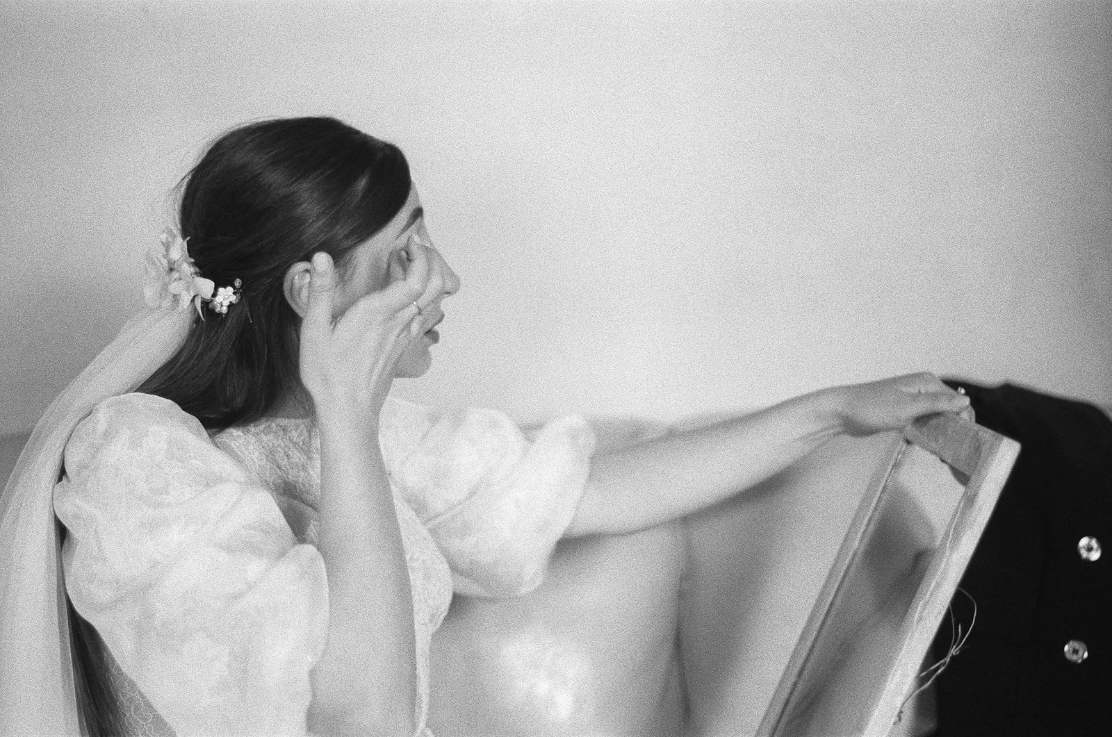 A woman with long hair, adorned with flowers, is in bridal attire and looks into a mirror while holding her face with one hand.