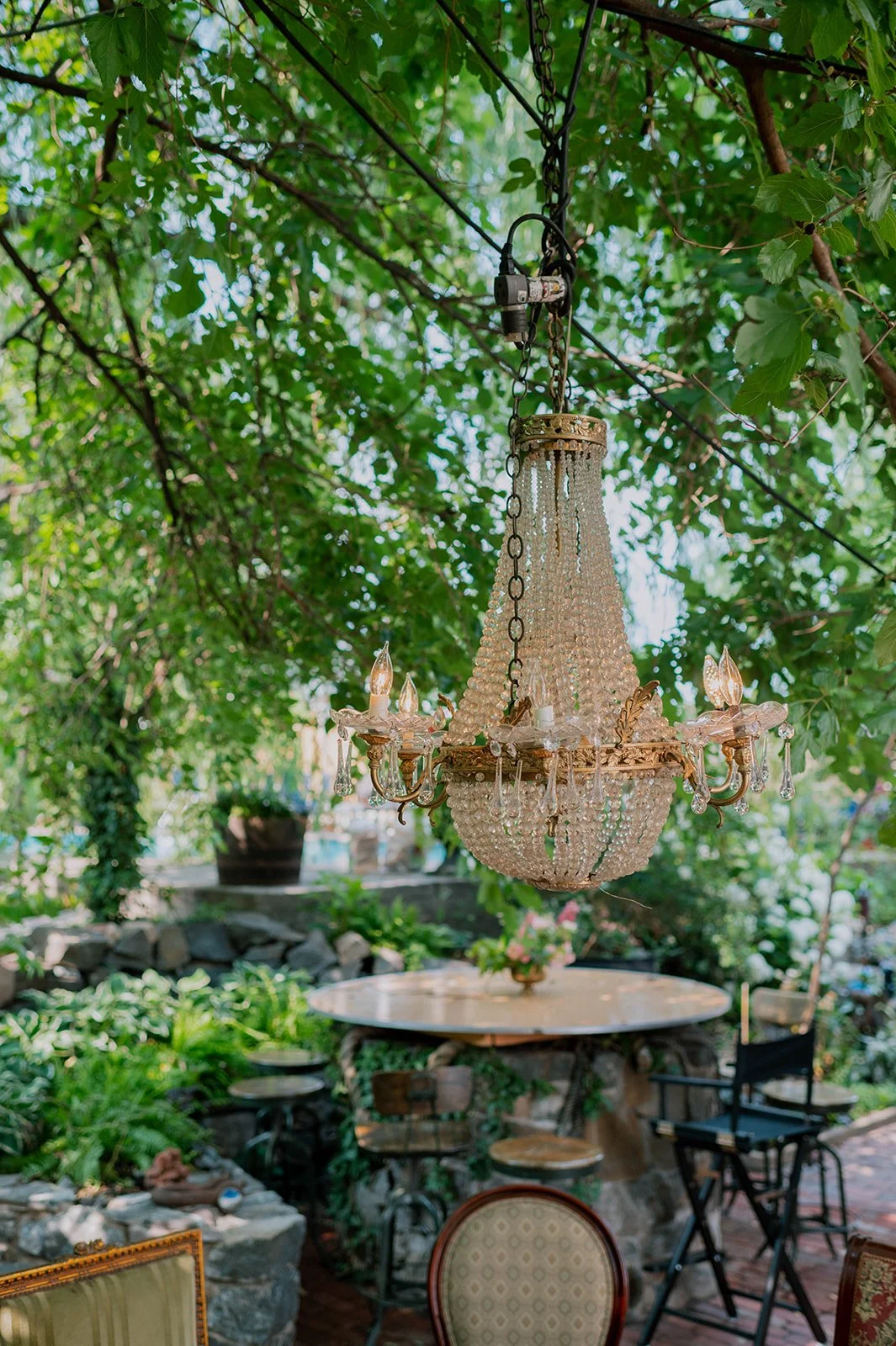 An ornate chandelier hanging outdoors among green leaves above a patio with a round table and chairs.