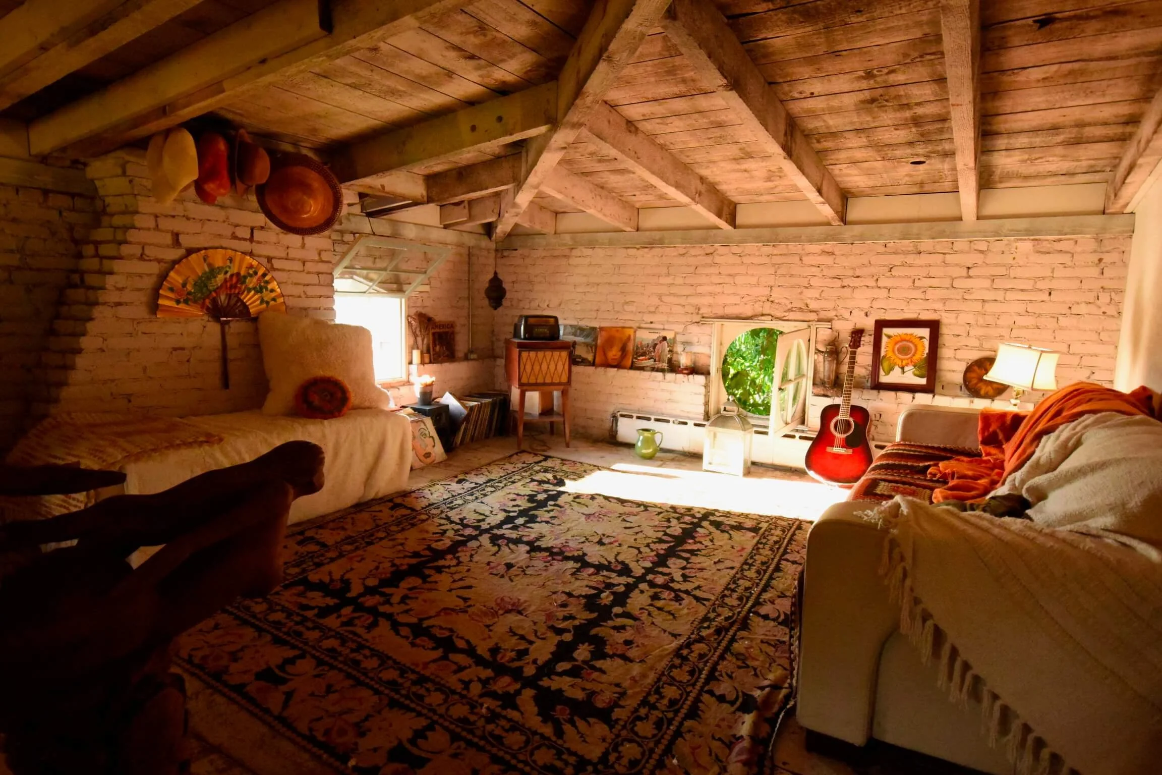 A cozy rustic living room with brick walls, wooden ceiling beams, a large ornate rug, and various furniture including sofas, a chair, and a guitar near a small window with greenery outside.