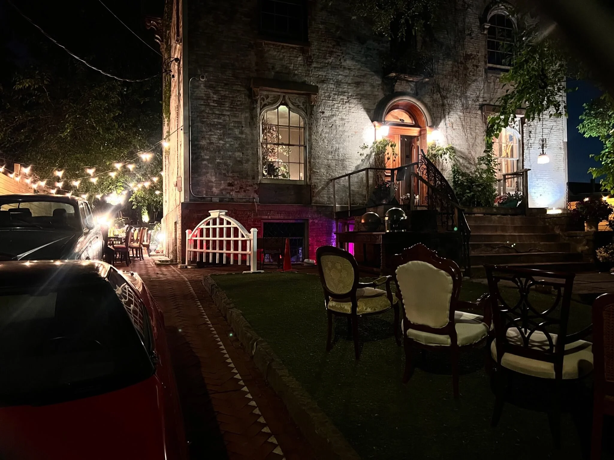 A nighttime scene of a brick house with a wooden door, decorated with outdoor string lights, patio furniture, and surrounding greenery.