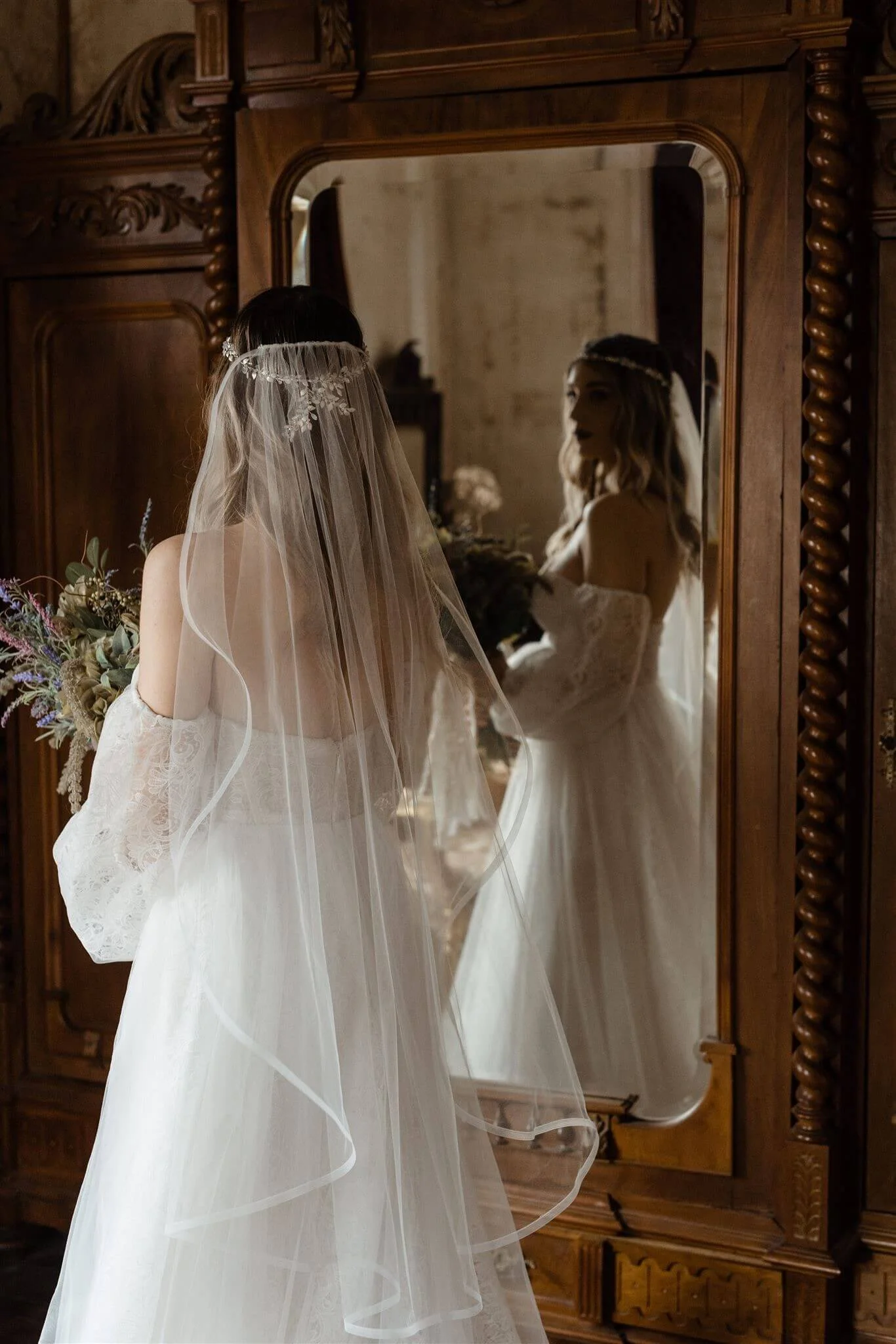 Bride wearing a lace wedding dress and veil, holding a bouquet, standing in front of a large wooden mirror in a vintage-style room.