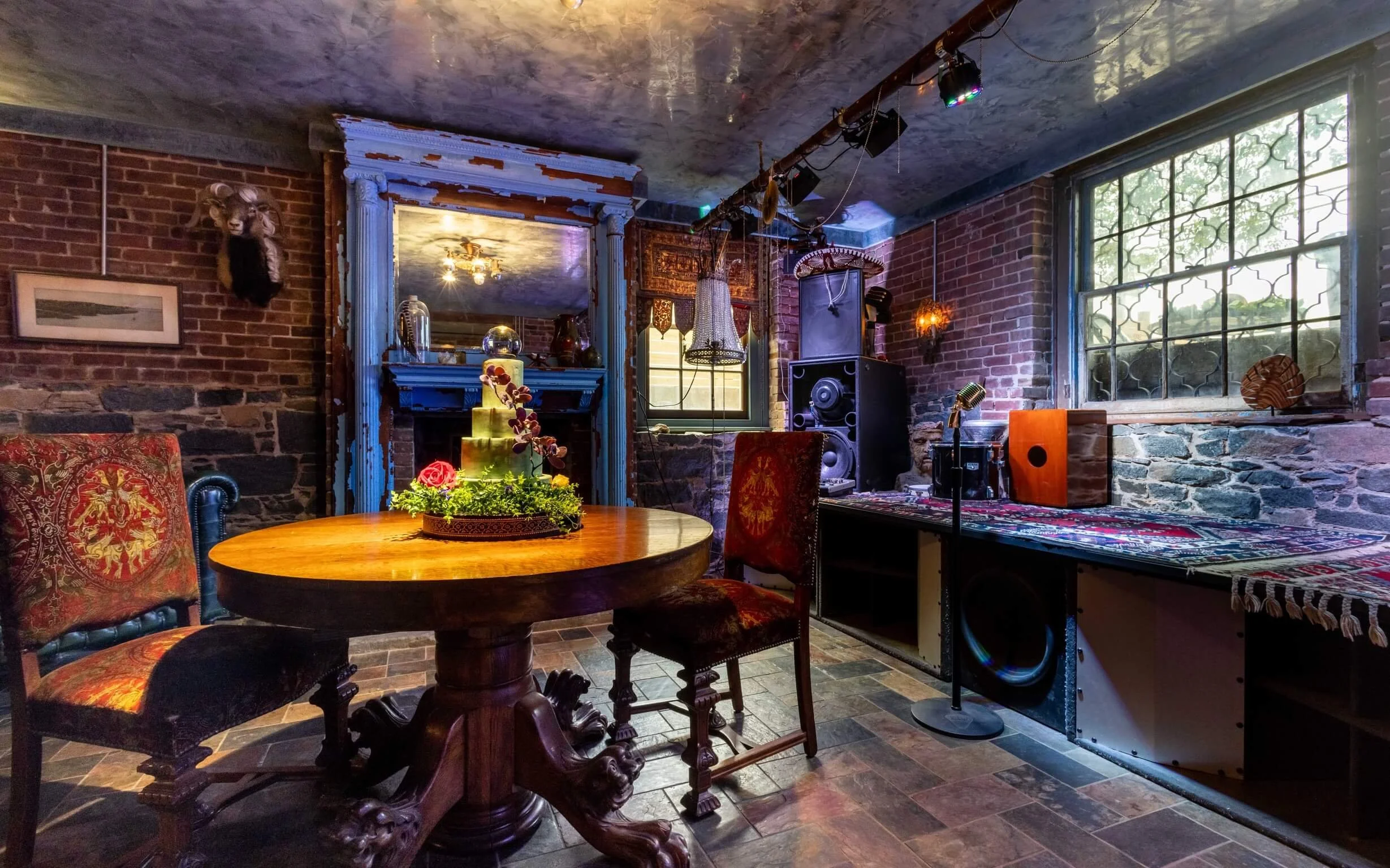 Room with brick walls, a round wooden table with floral centerpiece, vintage chairs, colorful lighting, a fireplace with blue trim, window with greenery outside, and audio equipment.