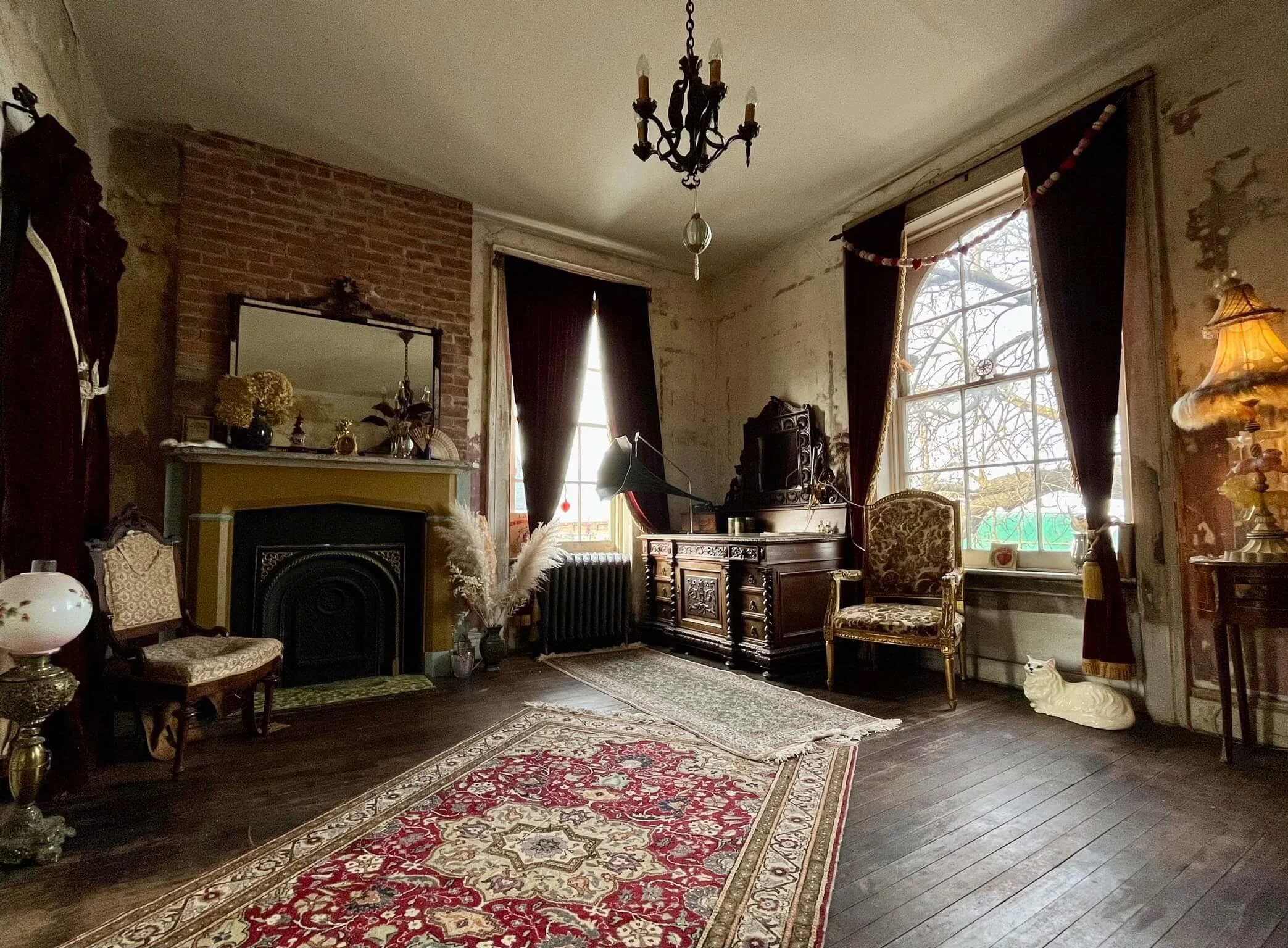 Vintage living room with exposed brick and plaster walls, hardwood floors, antique furniture, a patterned rug, a chandelier, and large windows with dark curtains.