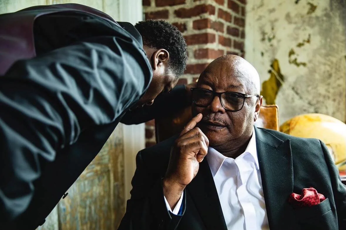 A man with glasses in a suit sitting at a desk with his hand near his chin, listening to a young man leaning in and speaking closely to him, in a room with exposed brick wall and rustic decor.