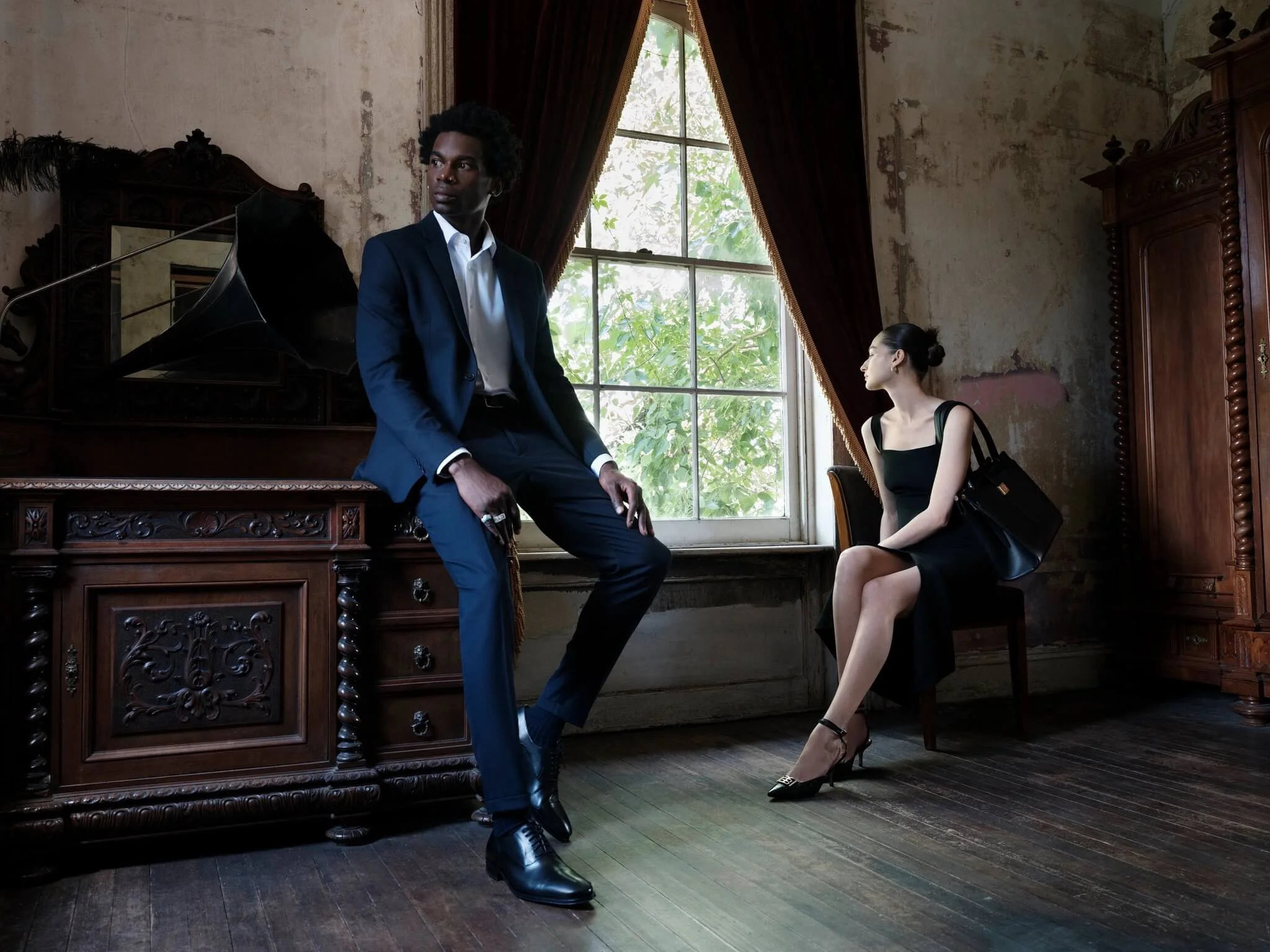 A man in a navy suit and a woman in a black dress sitting in a vintage room with an old wooden cabinet and a window with red curtains, with a black umbrella leaning against the vintage wooden cabinet.