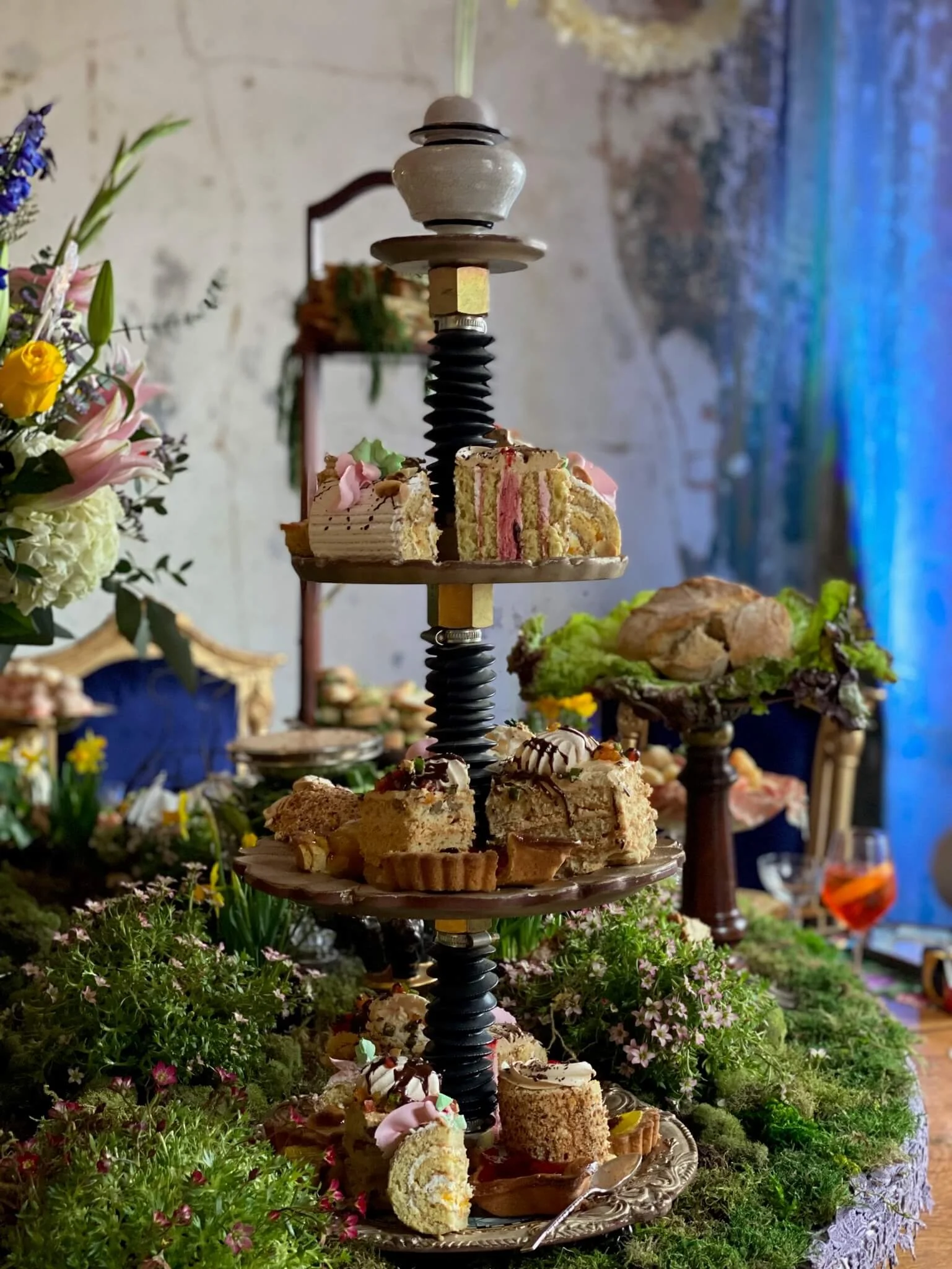A three-tiered dessert stand with assorted cakes and pastries, surrounded by flowers and greenery indoors.