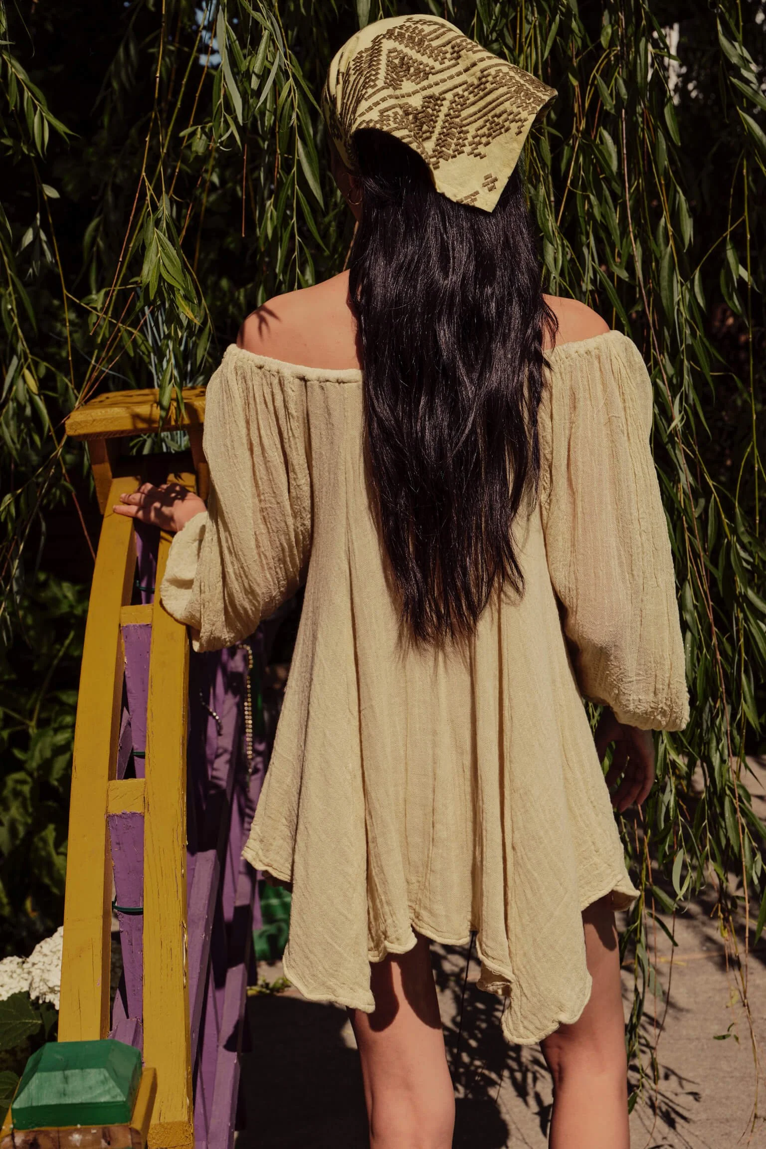 A woman with long dark hair wearing an off-shoulder beige dress and a patterned headscarf, standing outdoors near green leafy trees and a yellow wooden fence.