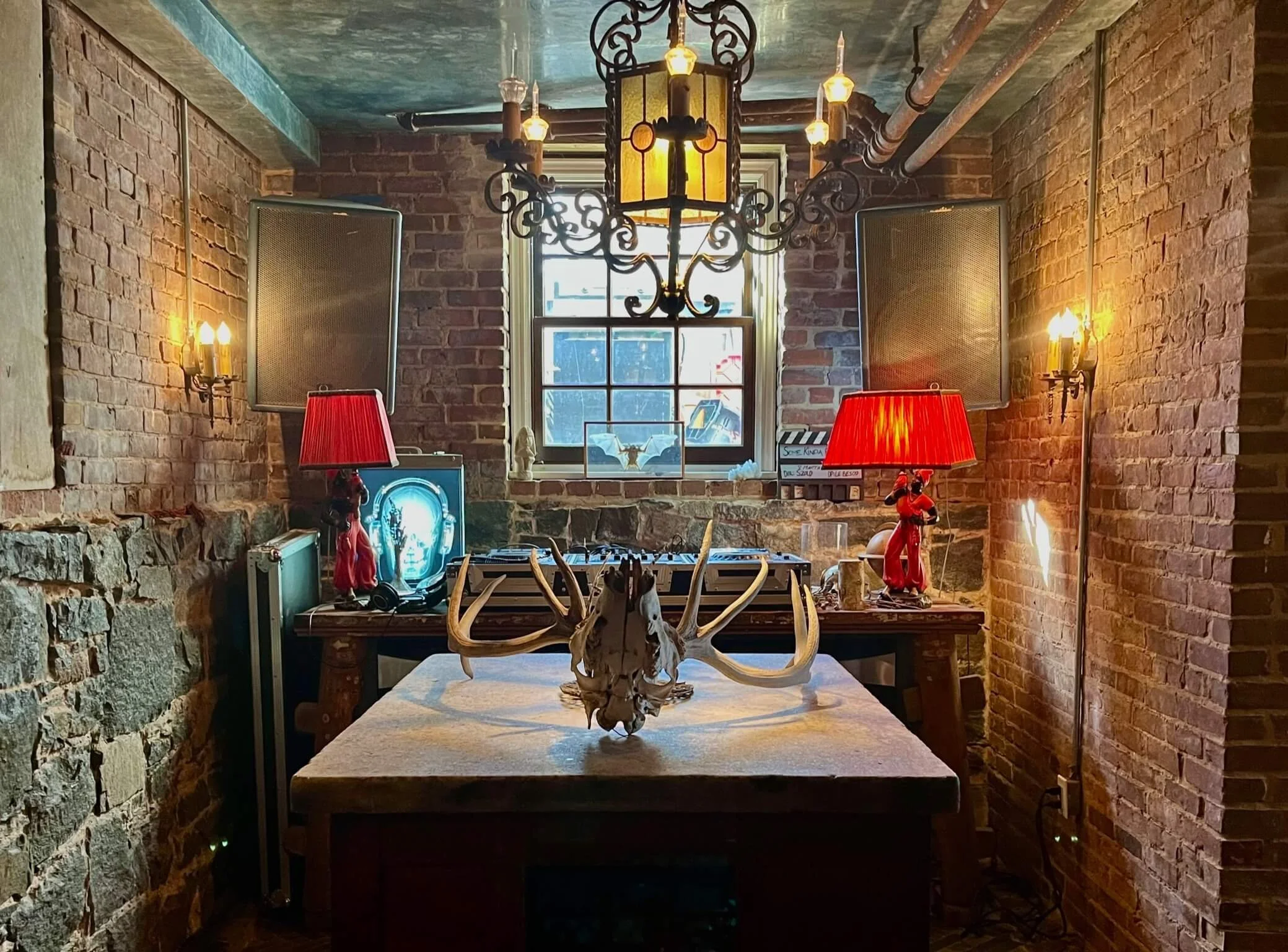 A rustic room with brick walls, a window, and a chandelier hanging from the ceiling. A table with a large animal skull with antlers is in the foreground. In the background, there are two red lamps, stereo speakers, a DJ mixing console, and decorative