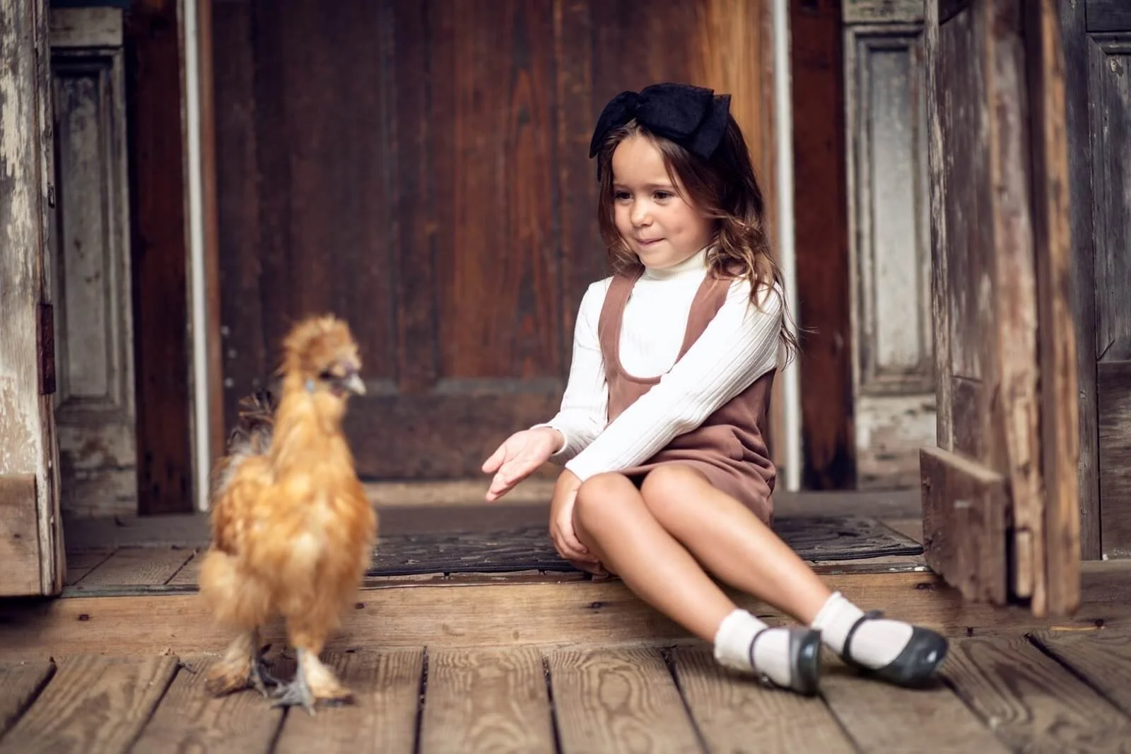 A young girl sitting on a wooden porch, reaching out to a small chicken in front of her.