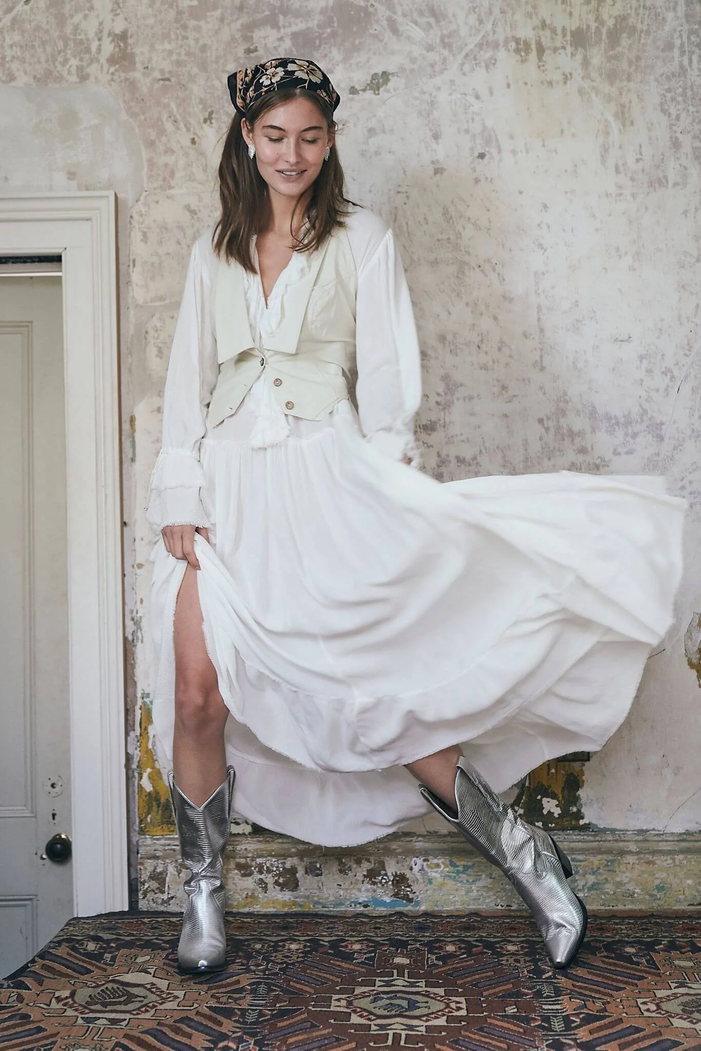 A woman in a white dress and silver cowboy boots standing on a patterned rug, holding up her skirt with one hand while looking down and smiling, with a distressed wall background.