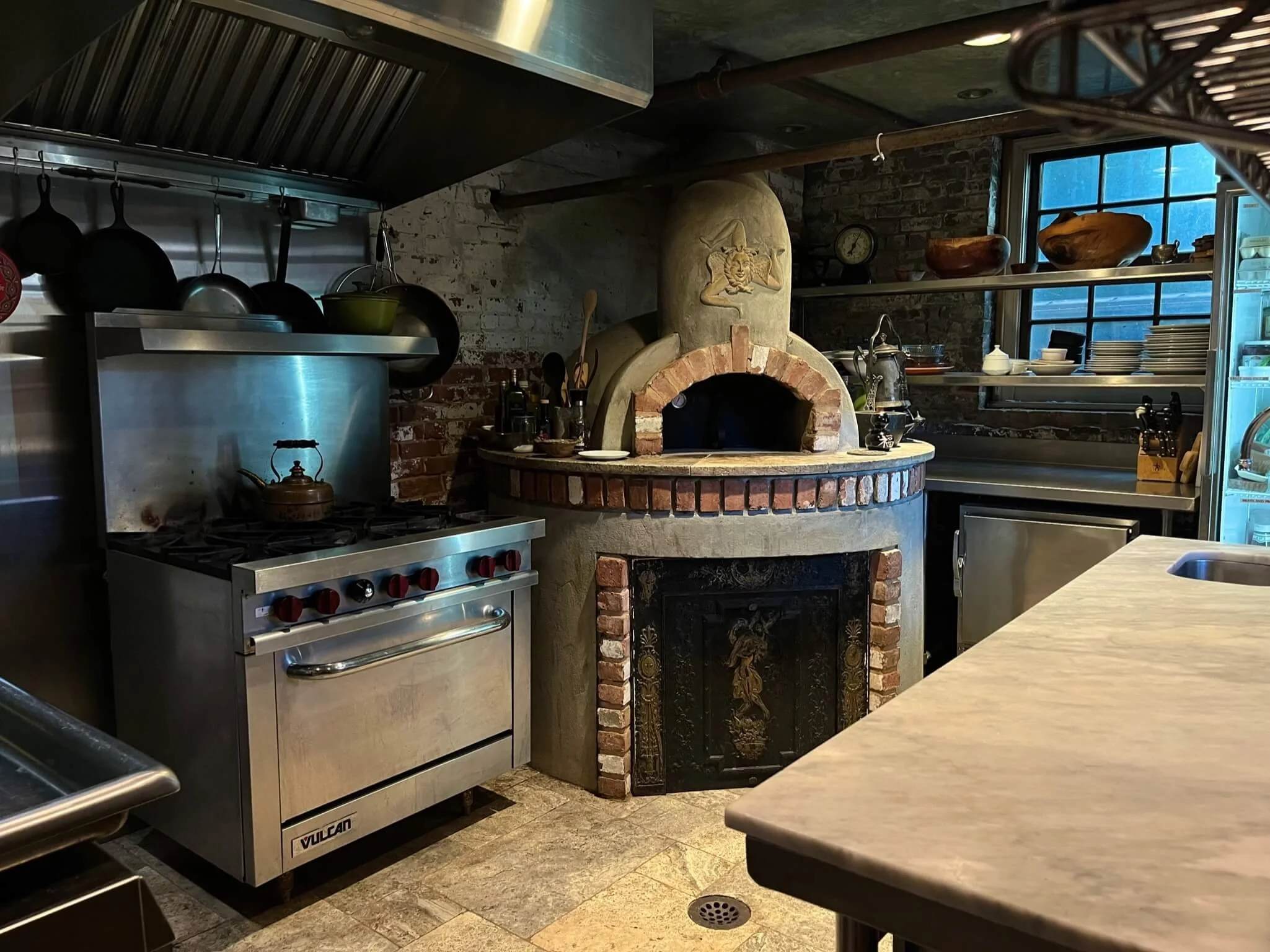A rustic kitchen with a wood-fired brick oven, stainless steel stove, open shelving with dishes and bowls, and a marble countertop.