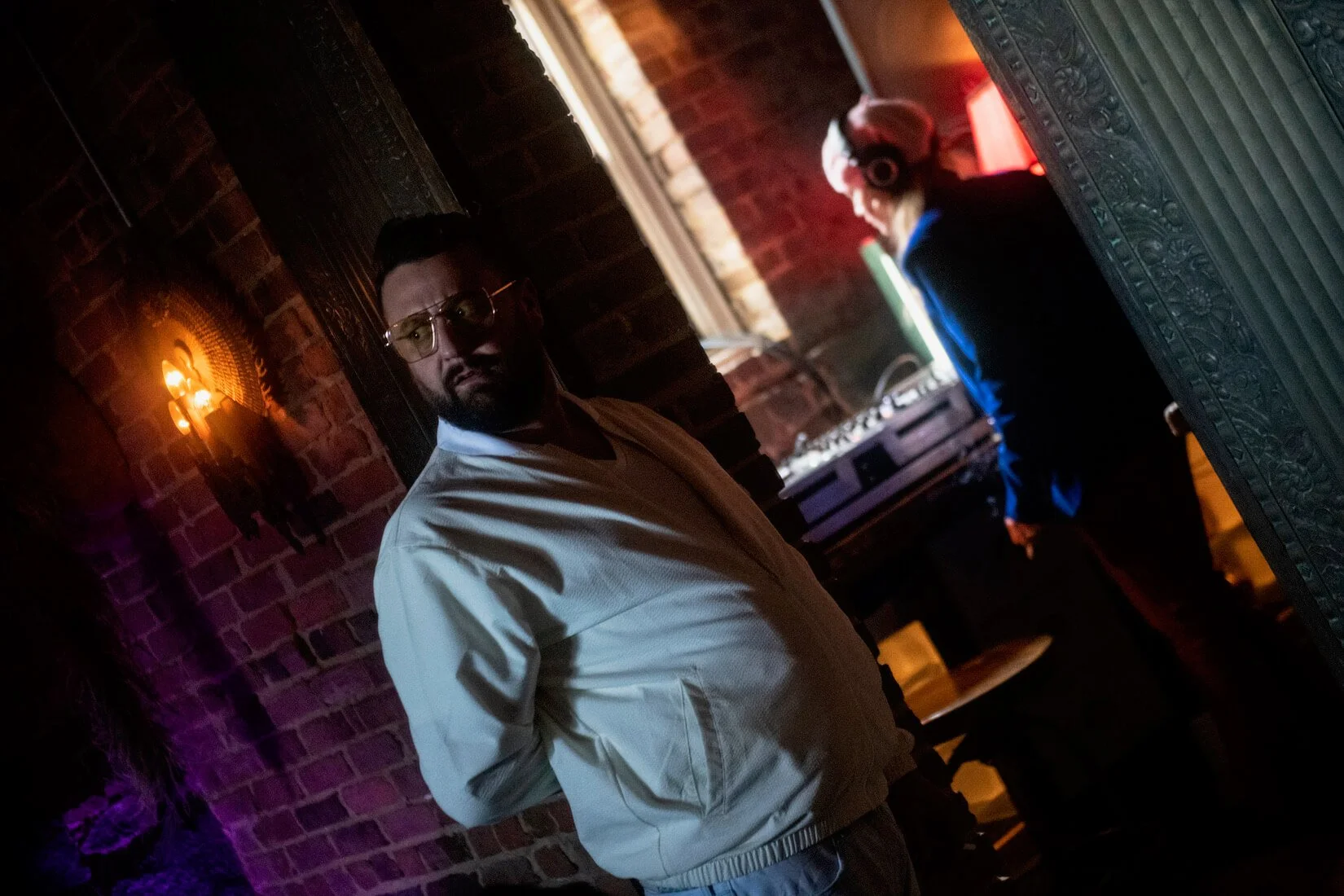 A man with glasses and a beard stands in a dimly lit room with exposed brick walls, wearing a light-colored jacket. In the background, a DJ wearing headphones works at a turntable near a window with natural light coming through.