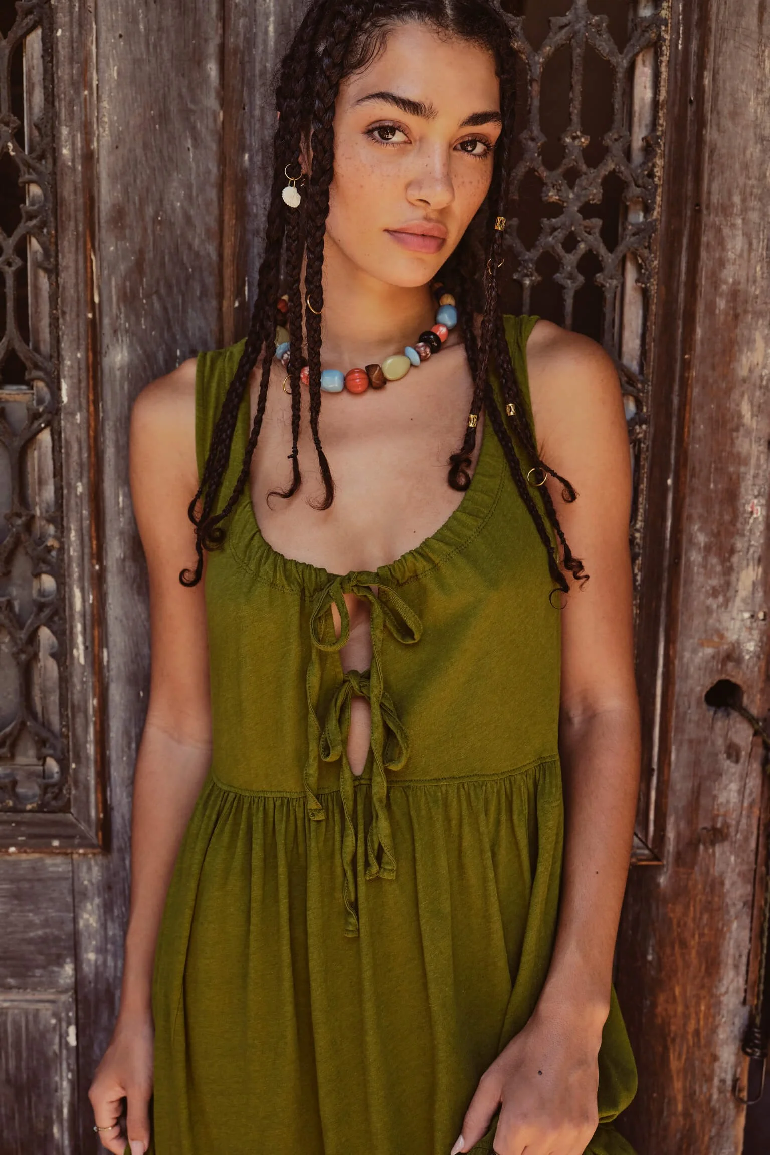 A woman with dark braided hair, wearing a green dress with a tie-front detail, and colorful beaded necklace, standing against a weathered wooden door.