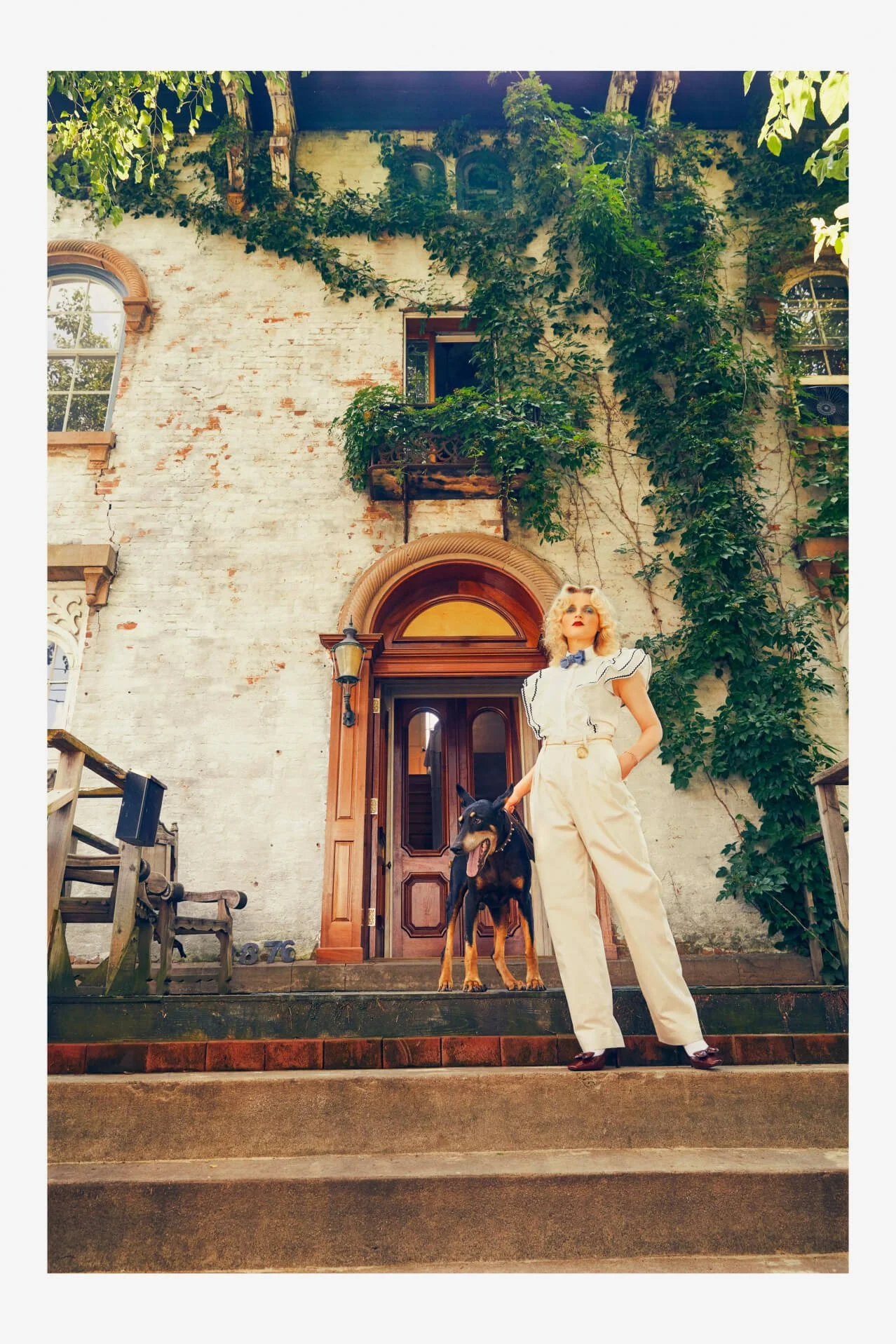 A woman standing on the steps of a vintage building with a black and tan dog. The building has a brick facade, wooden door and climbing ivy. The woman is wearing a white ruffled blouse and cream pants, with sunglasses and red lipstick.
