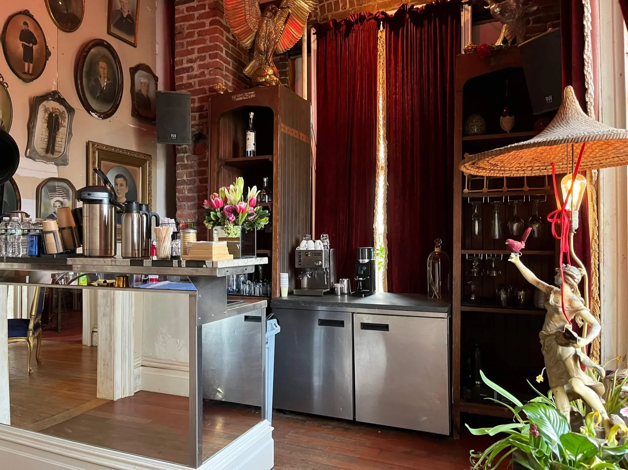 A cozy cafe or bar with a stainless steel counter, flowers, a coffee maker, and bottles. The background features framed portraits and a brick wall. There is a shelf with glasses and decorative items, and a table with water bottles and cups. A statue 
