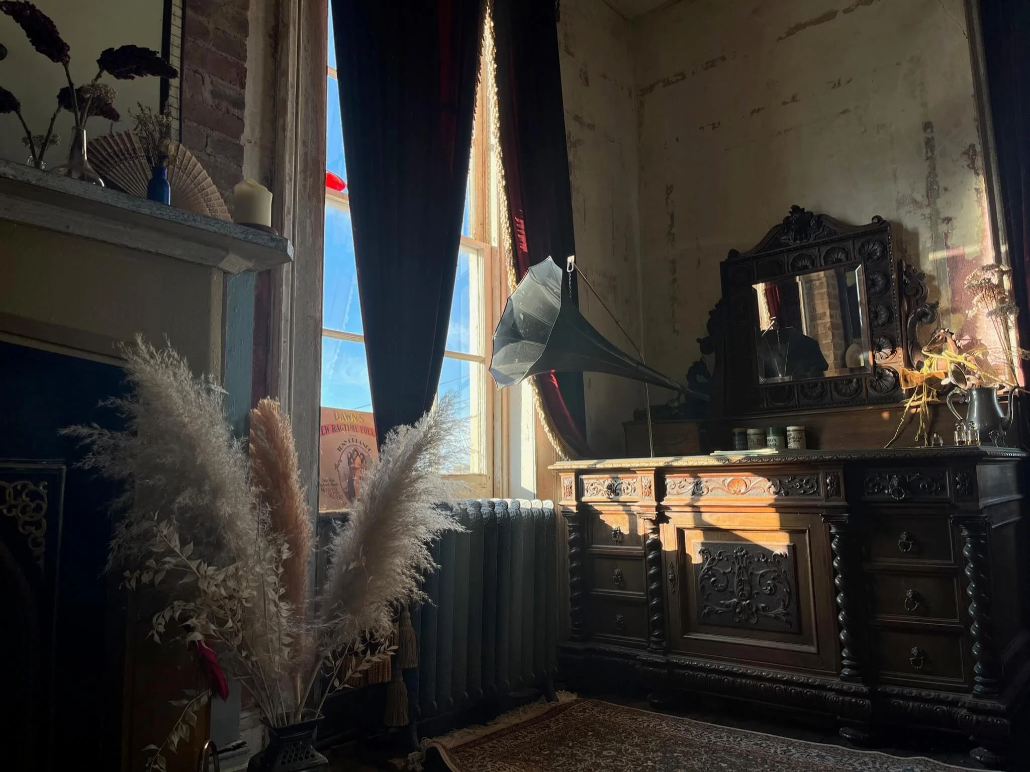 Vintage interior room with an ornate wooden dresser and mirror, a large window with dark curtains, an old gramophone, dried pampas grass in a vase, and old-fashioned decor. Sunlight streams through the window.