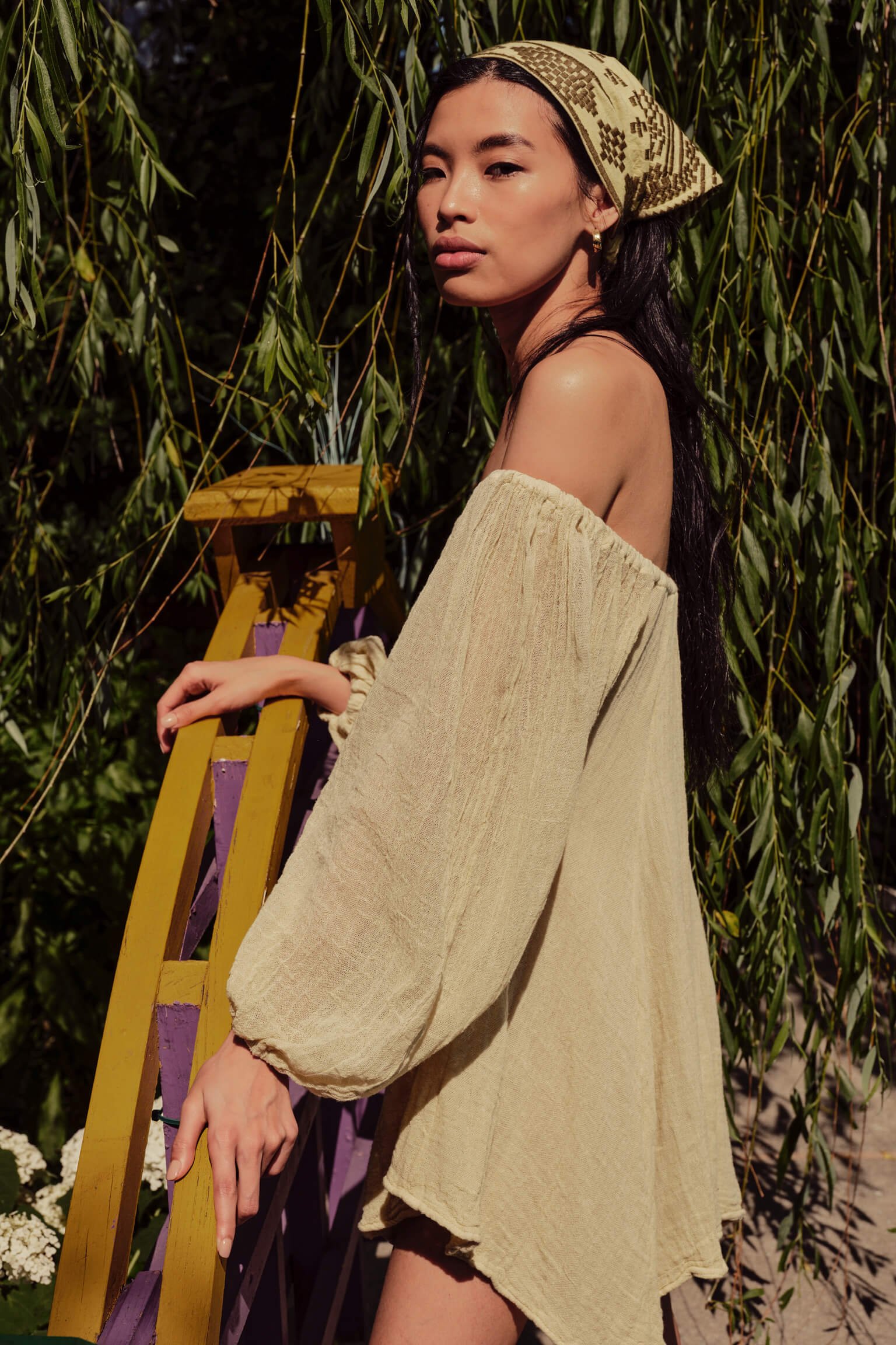 A woman with long dark hair wearing a beige off-shoulder dress and a patterned headscarf stands outdoors near a colorful yellow and purple ladder with hanging green foliage behind her.