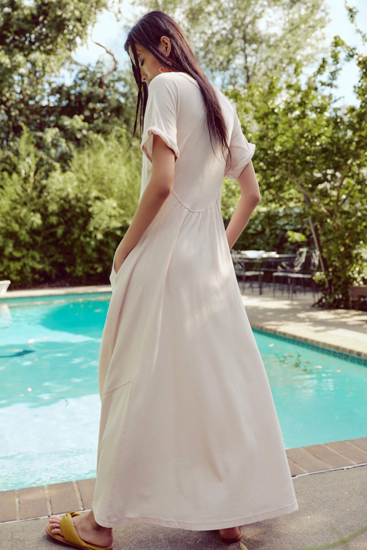 A woman in a light-colored dress standing near a swimming pool, with trees in the background.