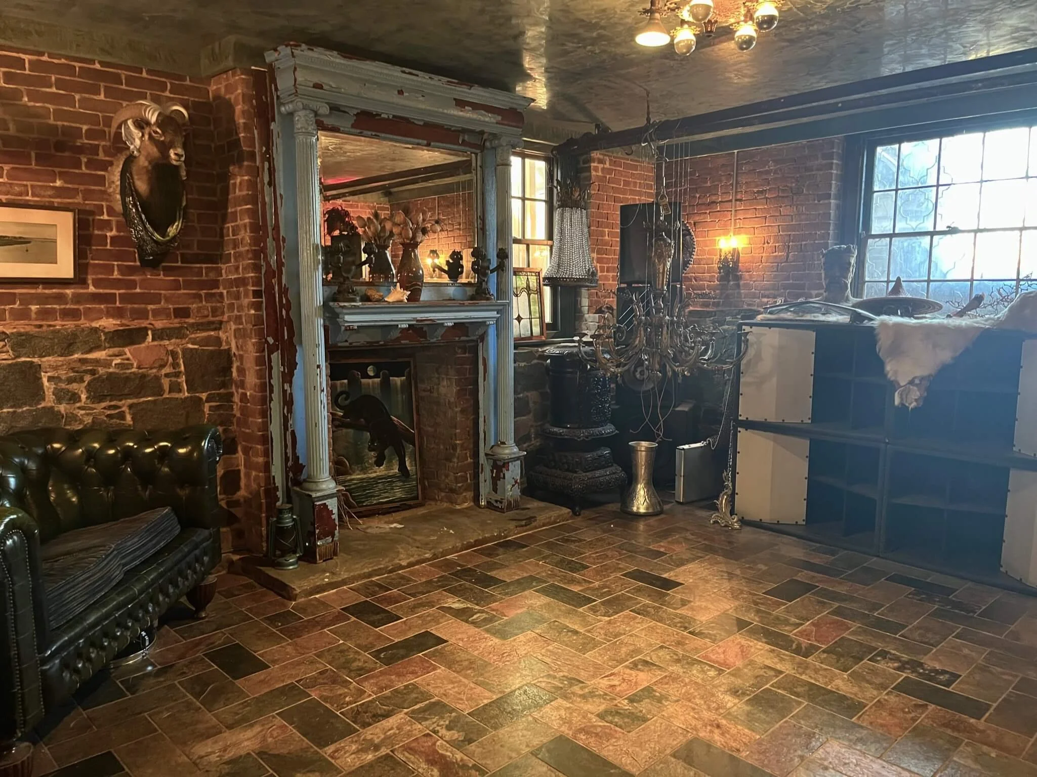Interior of a room with brick walls and a tiled floor, decorated with various antiques, sculptures, and furniture, including a chandelier, a mounted animal head, and a leather sofa.