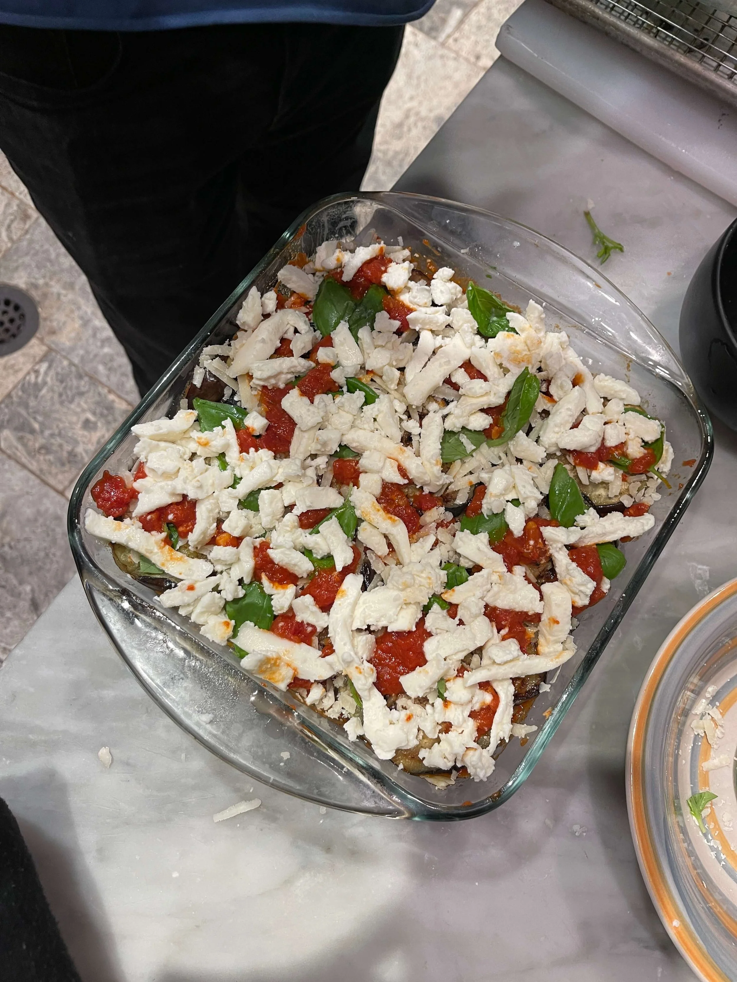 Layered eggplant dish topped with chopped mozzarella cheese, fresh basil leaves, and marinara sauce in a glass baking dish.