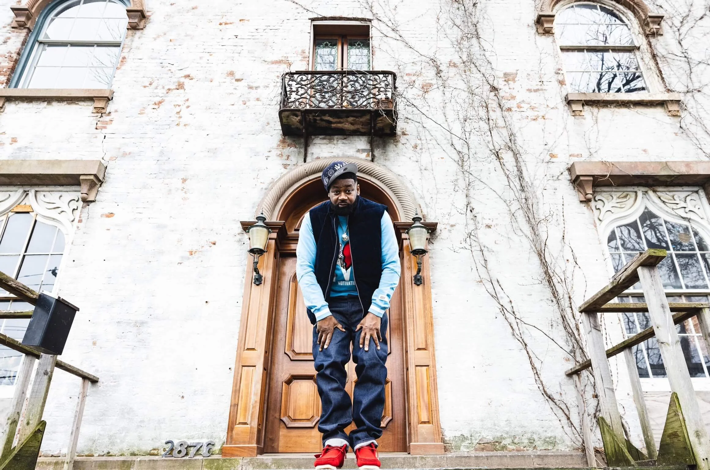 A man standing on stairs in front of a wooden door with arched frames, on a white brick building with ivy vines, two lanterns flanking the door, and windows with decorative trim, with house number 2876 visible at the bottom.