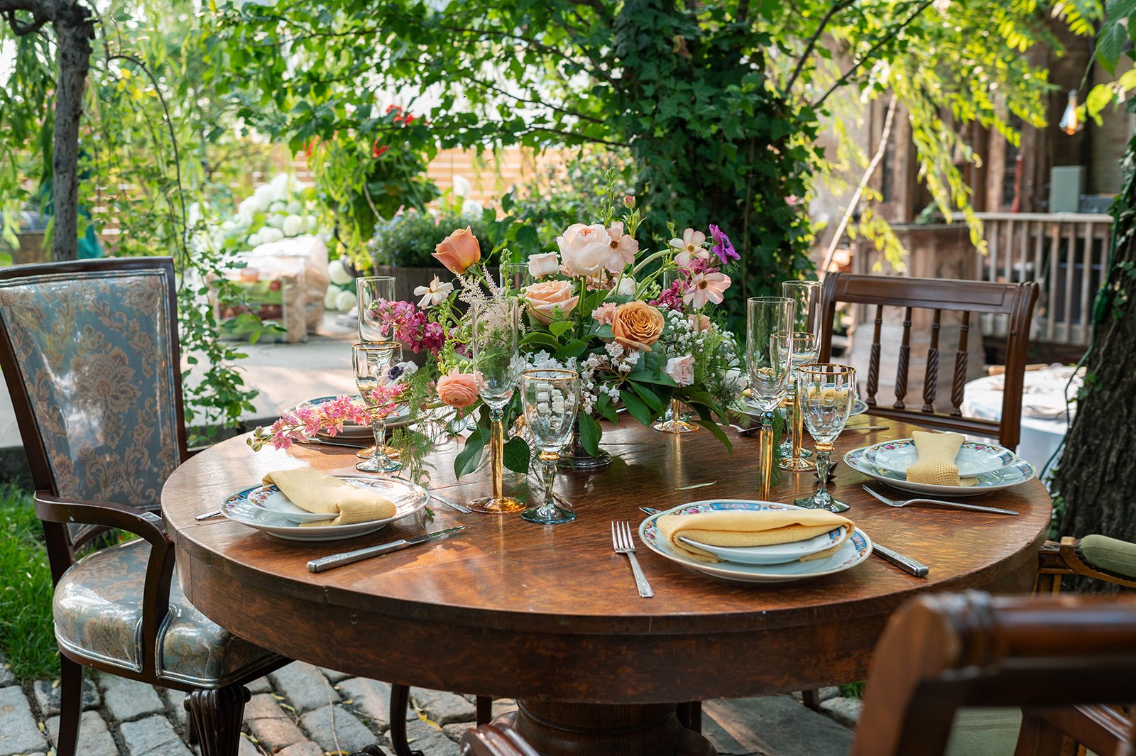 An outdoor dining table set with a floral centerpiece, plates, napkins, silverware, and glassware, surrounded by chairs, in a garden-like setting with trees and greenery.