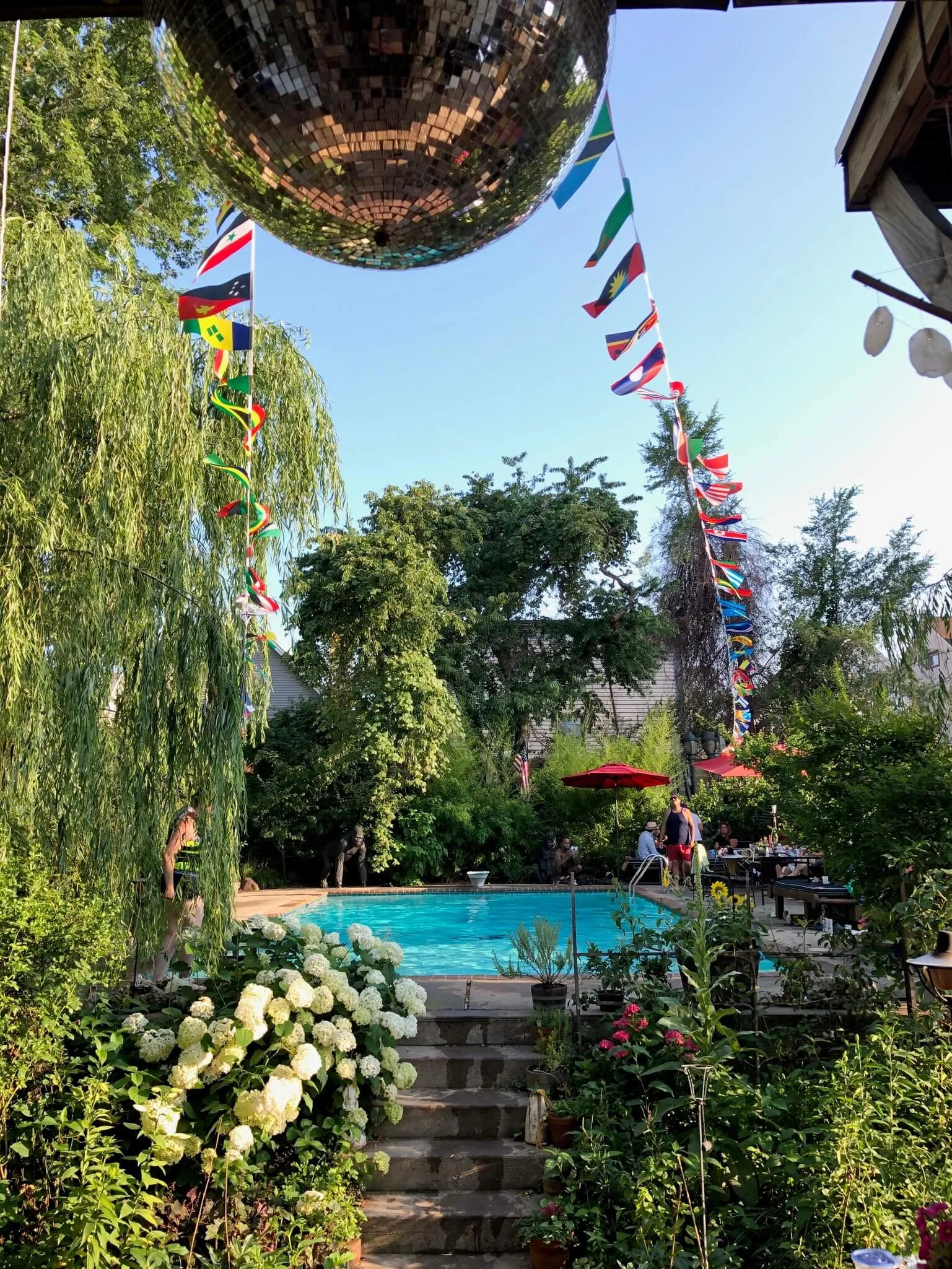 A backyard with a swimming pool surrounded by lush greenery, flowers, and trees. There are string flags of various countries hanging above and a large disco ball reflecting light. People are gathered around the pool area, some sitting under red umbre