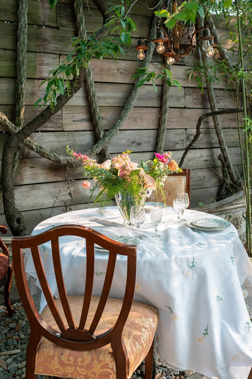 A round table with a white embroidered tablecloth set outdoors against a wooden fence with climbing plants. The table features a large floral centerpiece with pink and peach flowers in a glass vase, surrounded by crystal glasses and plates. Wooden ch