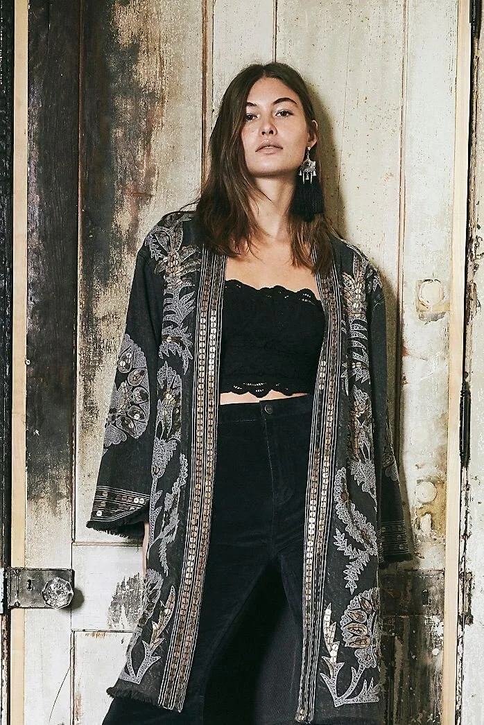 Young woman with long dark hair standing against a rustic wooden wall wearing a black lace crop top and a long, embroidered black kimono-style jacket.